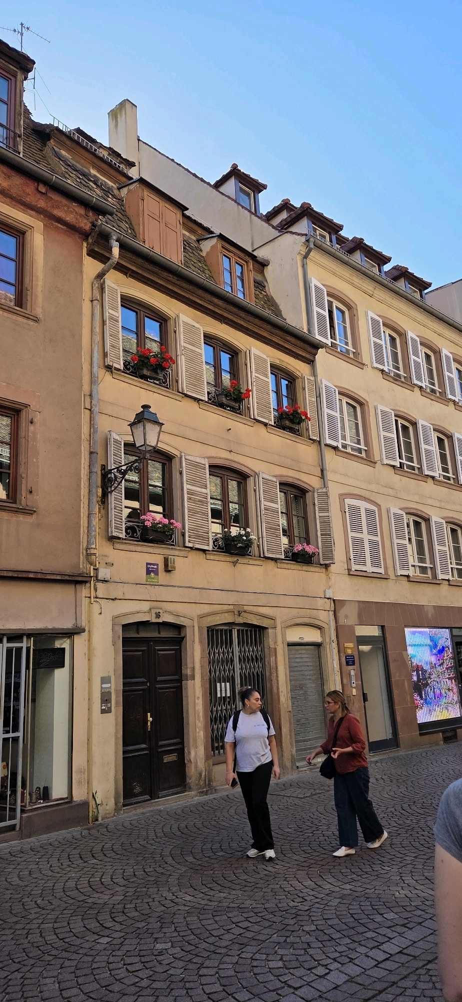 The image captures a street scene in Strasbourg, France, characterized by its historic architecture. The buildings lining the cobblestone street are multi-story structures with distinct window designs, many featuring white louvered shutters and window boxes filled with red and pink flowers. The facades are painted in warm earthy tones, such as beige and terracotta.
Two women are the primary human subjects in the foreground. The woman on the left, wearing a light gray t-shirt and black pants, is walking towards the right side of the frame, looking slightly to her left. She has dark hair pulled back and is carrying a backpack. The woman on the right, dressed in a rust-colored top and dark blue pants, is walking in the same direction, looking ahead. She has blonde hair and a dark shoulder bag.
On the left side of the street, a shop window is visible, displaying items for sale. Further down the street, a large, dark wooden double door is set within an arched opening, marked with the number 16. To its right, a metal gate is closed over another storefront. The buildings extend upward, showing multiple levels of windows and sloped rooftops with dormer windows. The sky above is clear and blue. The pavement is made of dark, circular cobblestones laid in a fan pattern.