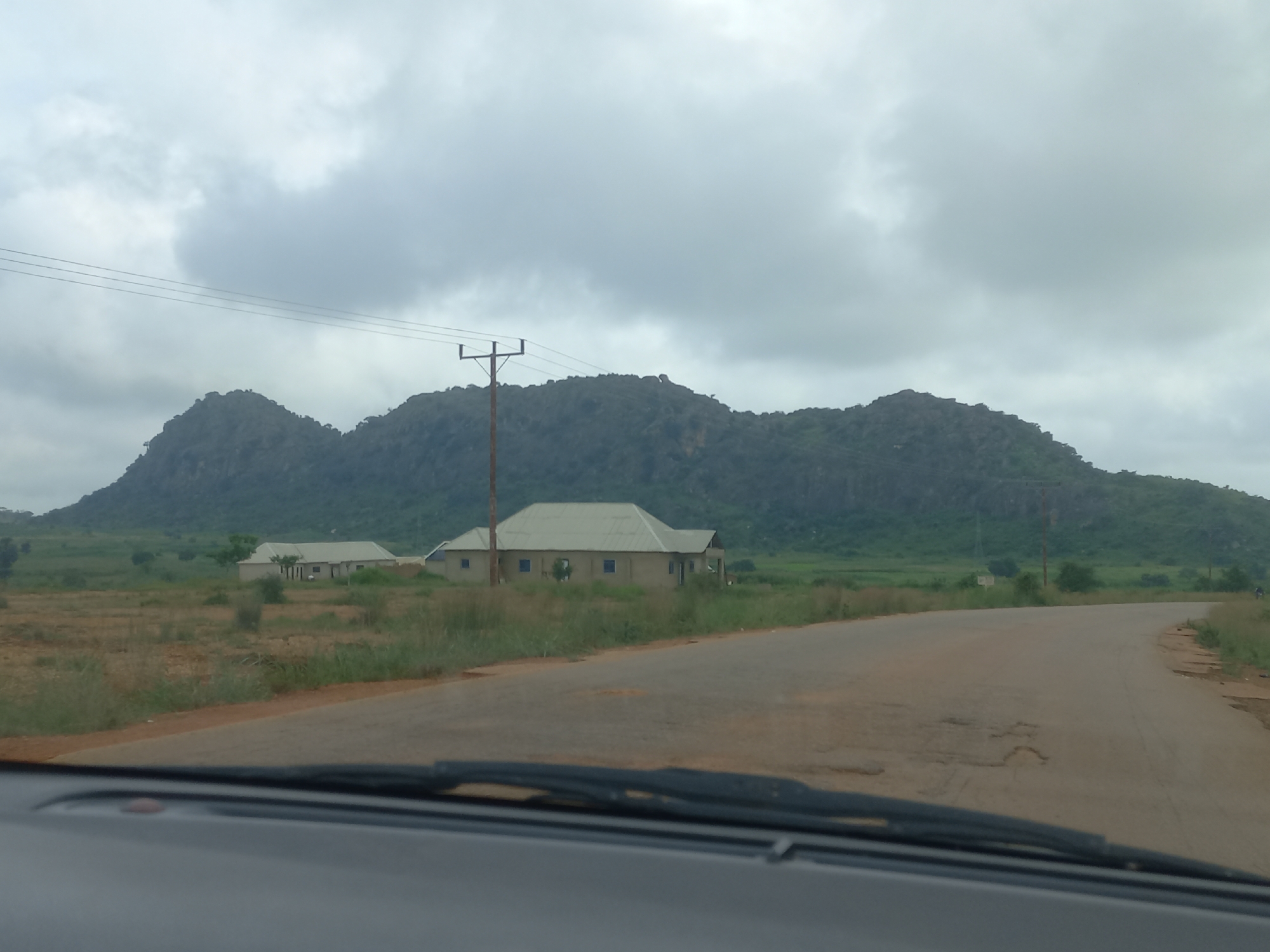 The image shows a wide, paved road curving to the right. The road surface is weathered and shows several potholes. On the left side of the road, there are two buildings with light-colored walls and light-colored roofs. The foreground is dominated by the dashboard and windshield of a vehicle. In the background, a large, rocky mountain range rises under a cloudy sky. Electrical poles and wires are visible in the distance. The landscape appears to be rural, with some vegetation and scattered trees. The scene is likely located in the vicinity of Mangu Halle, Nigeria.