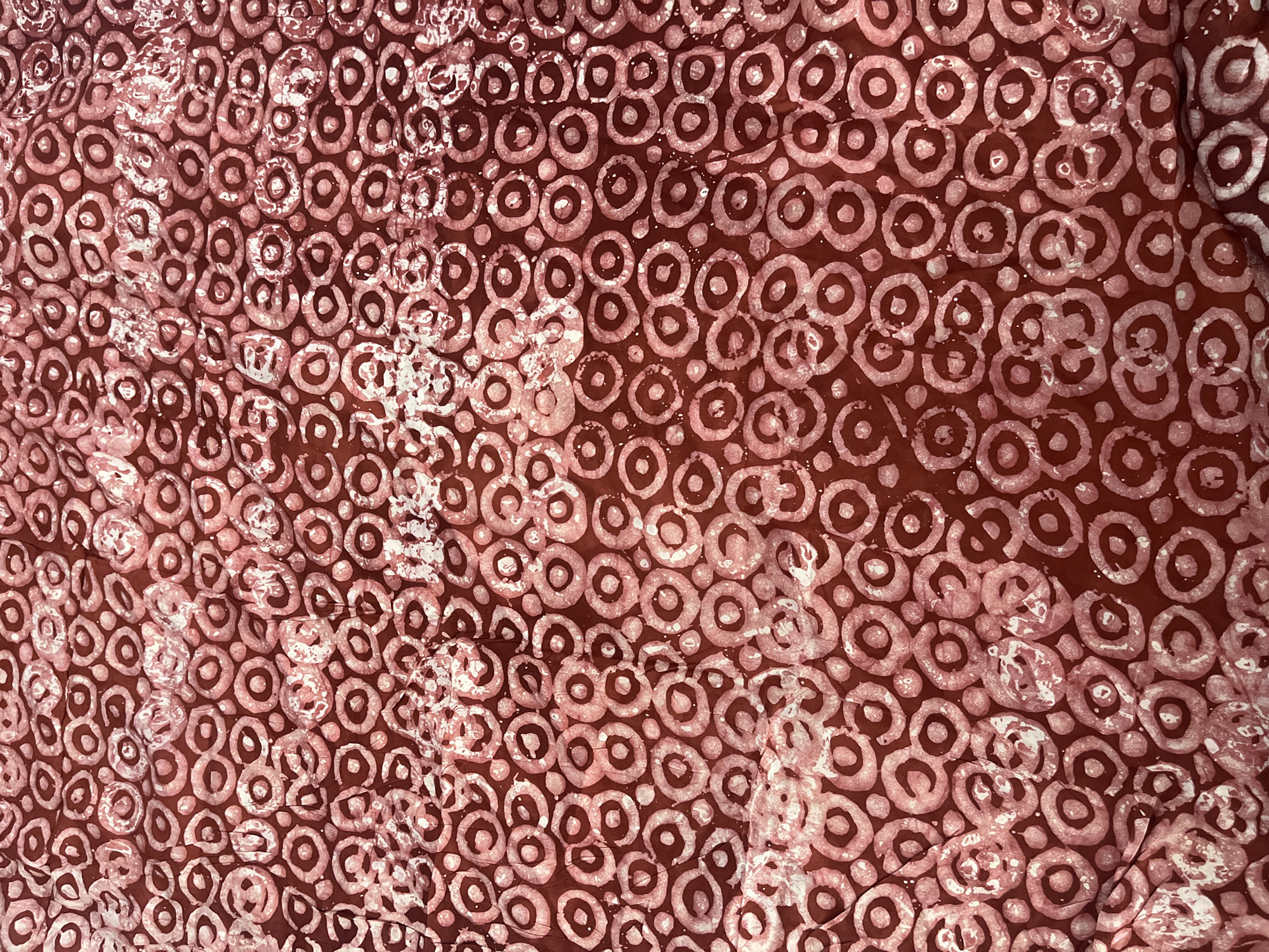 The image shows a close-up view of a patterned textile with a repeating design of circles or ring-like shapes. The circles appear in two tones, a darker reddish-brown and a lighter pinkish-white, suggesting a possible dyeing or printing technique. The fabric has a textured or slightly wrinkled appearance. There are no people, objects or actions to be seen in this image. The location of Jos, Nigeria cannot be determined from this picture.