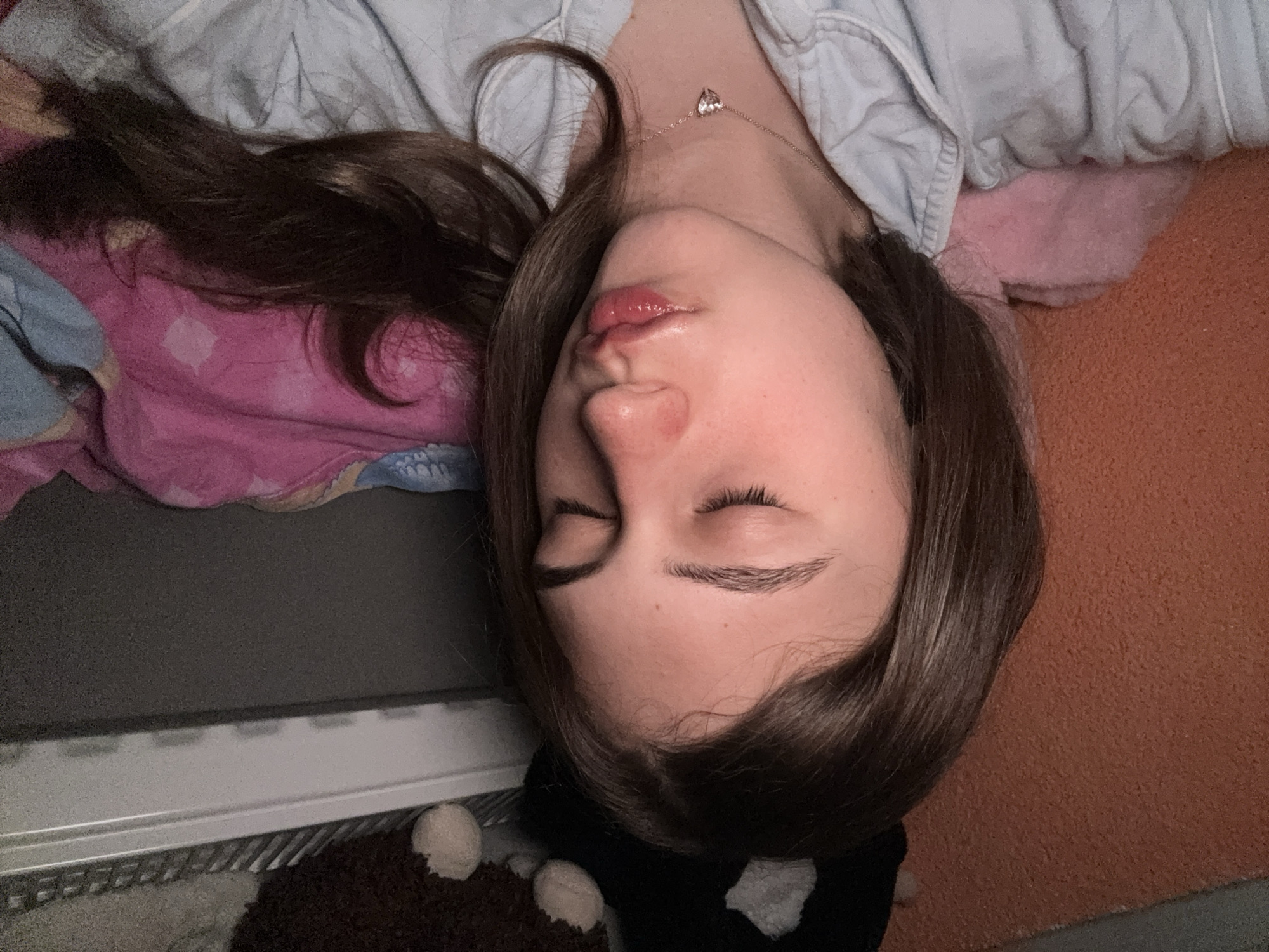 A young person with long brown hair is lying down with their eyes closed. They are wearing a light-colored shirt and a necklace with a heart pendant. The person is lying on a bed with various covers and pillows, including a pink one and one with a patterned design. The background also includes a heating radiator and what appears to be a stuffed animal. There is no evidence to determine if the image was taken in Panagyurishte, Bulgaria.
