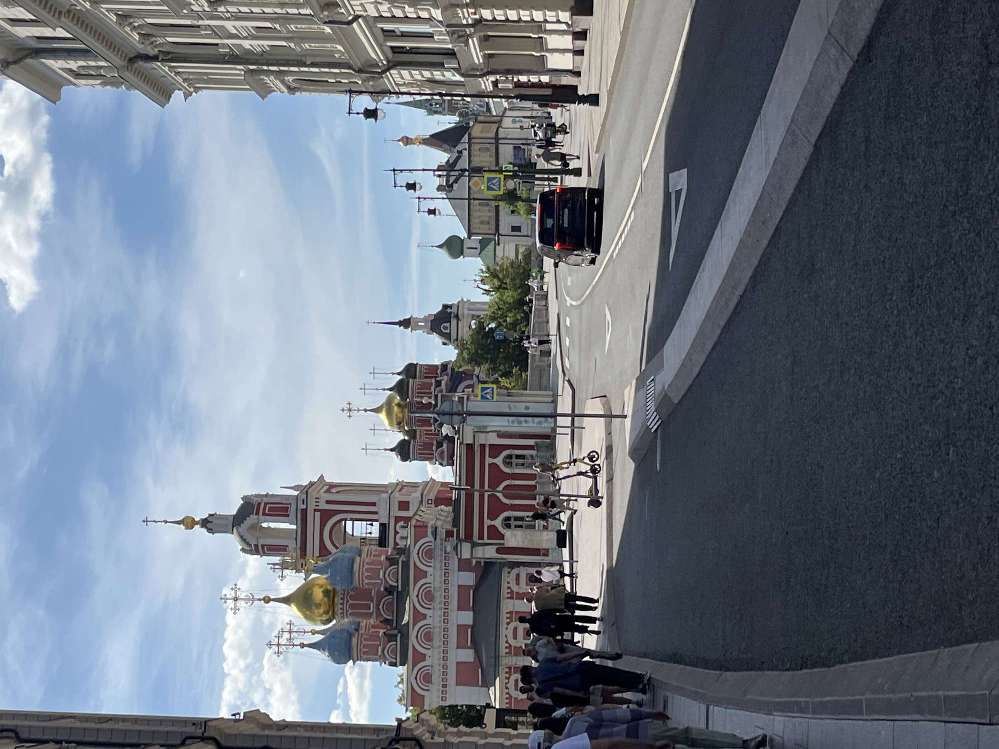In Moscow, Russia, a street scene unfolds with a prominent red and white domed church on the left, featuring gold and blue domes topped with crosses. A white building with a grand facade stands on the right. Pedestrians walk along the sidewalk on the left, and a dark-colored SUV drives down the asphalt road. Several streetlights line the right side of the road. The sky is a mix of blue with scattered white clouds. In the mid-ground, a yellow electric scooter is parked on the sidewalk. The road curves to the right, with white directional arrows painted on the asphalt.