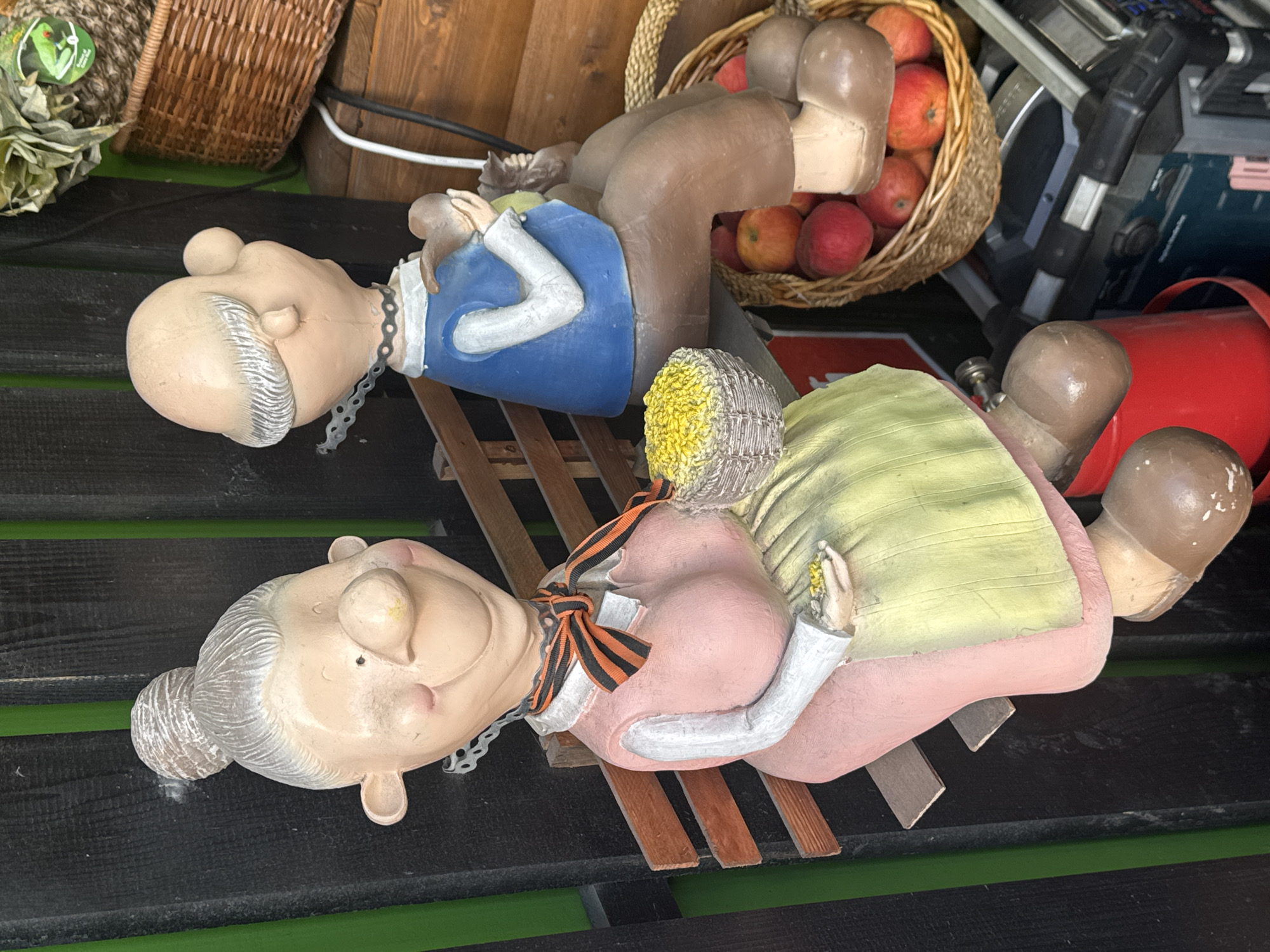 Two painted figurines of an elderly couple are reclining on a wooden bench. The male figurine is wearing a blue shirt, and the female figurine is wearing a pink dress and has a yellow head covering. An orange and black striped ribbon adorns her neck. In the background, there is a basket filled with apples. The scene appears to be outdoors, on a black surface with green accents, though the specific location in Saint Petersburg, Russia, is not discernible from the image.