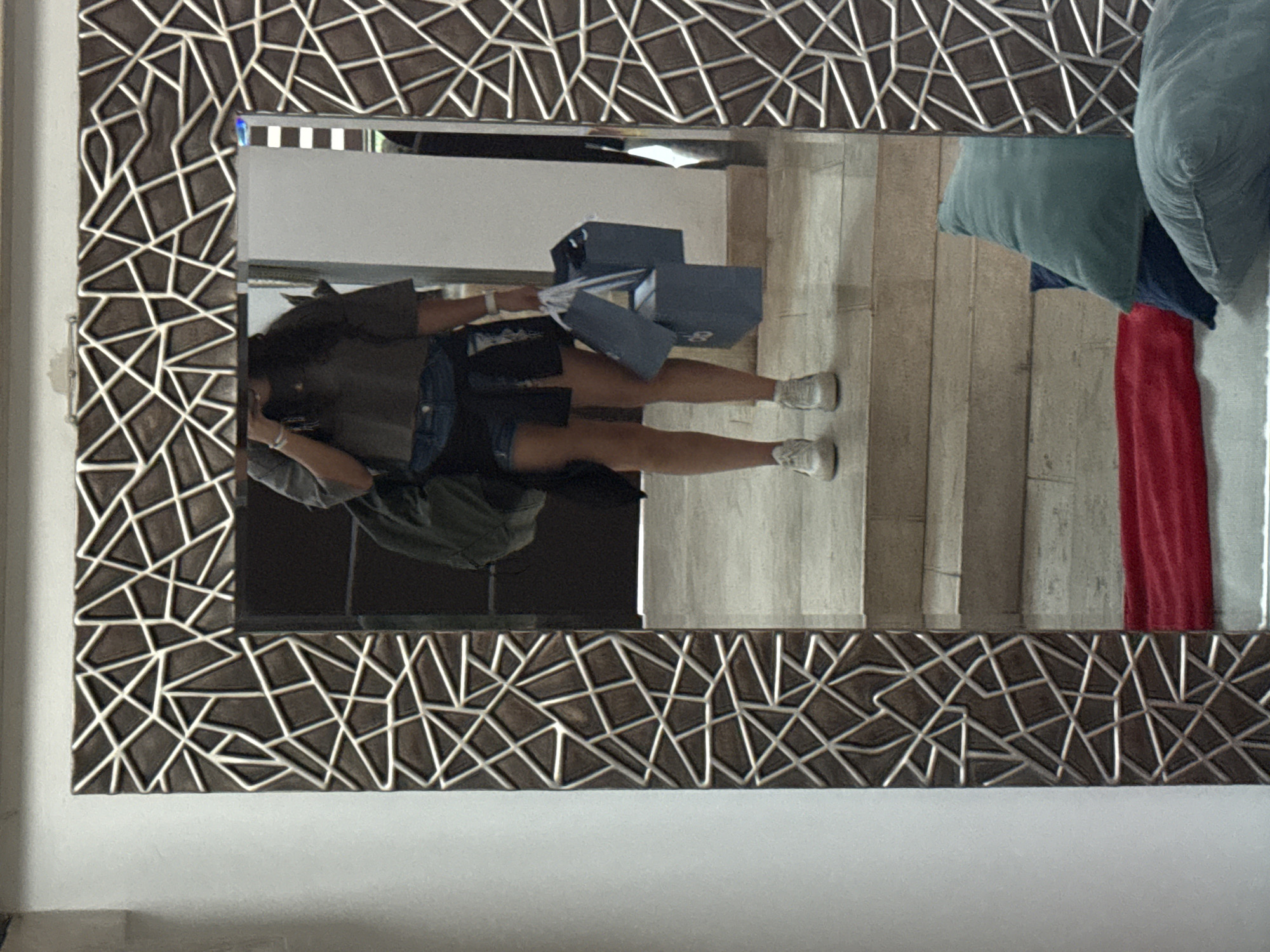 The image shows a mirror reflecting a person holding shopping bags. The person is wearing shorts and sneakers. Behind the person is a wall, and to the right of the reflection are pillows and a red item. The mirror has a patterned frame. There is no information to determine if the photo was taken in San Bernardino Tlaxcalancingo, Mexico.