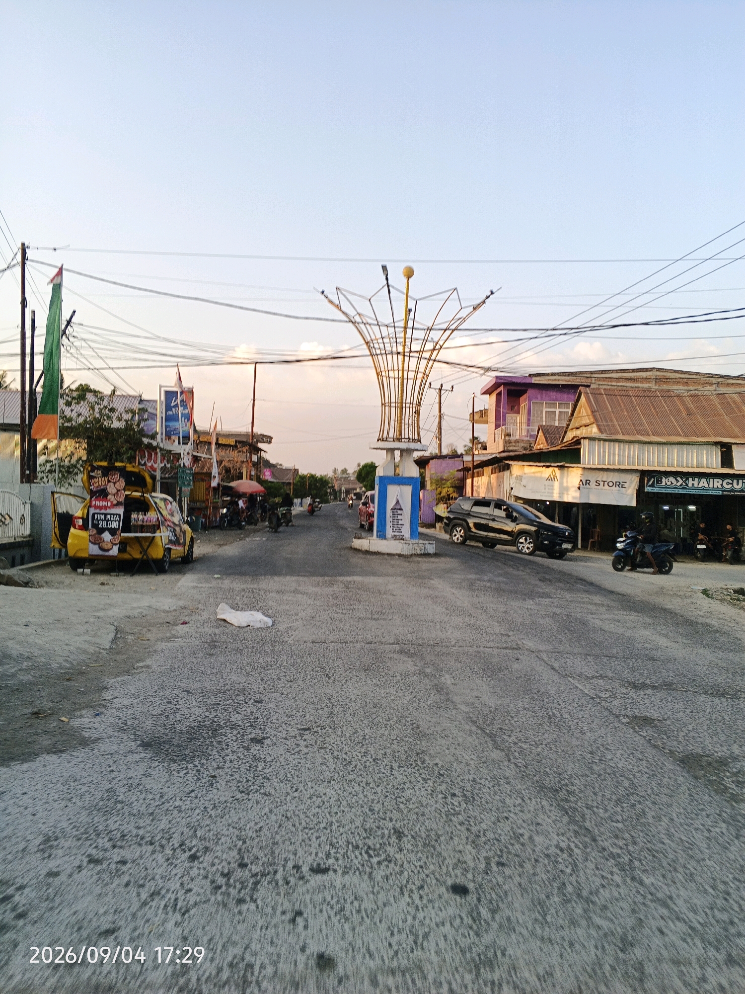 The image depicts a street scene in Bulu Cenrana, Indonesia, likely in the late afternoon given the lighting. The road surface is worn and shows patches of darker asphalt. On the left side of the road, a yellow car is parked with its trunk open, displaying what appears to be food items for sale with a "PROMO Rp 20.000" sign. Further down the street, there are various shops and businesses lining both sides. One building has a sign that reads "BOX HAIRCUT". Several motorcycles are visible, with one black SUV driving away from the camera towards the right. A decorative metal sculpture stands in the center of the road, positioned on a concrete base. The sky is clear and light blue with some wispy clouds. Utility poles and overhead wires are prominent throughout the scene. The overall impression is of a typical bustling street in a local Indonesian town.