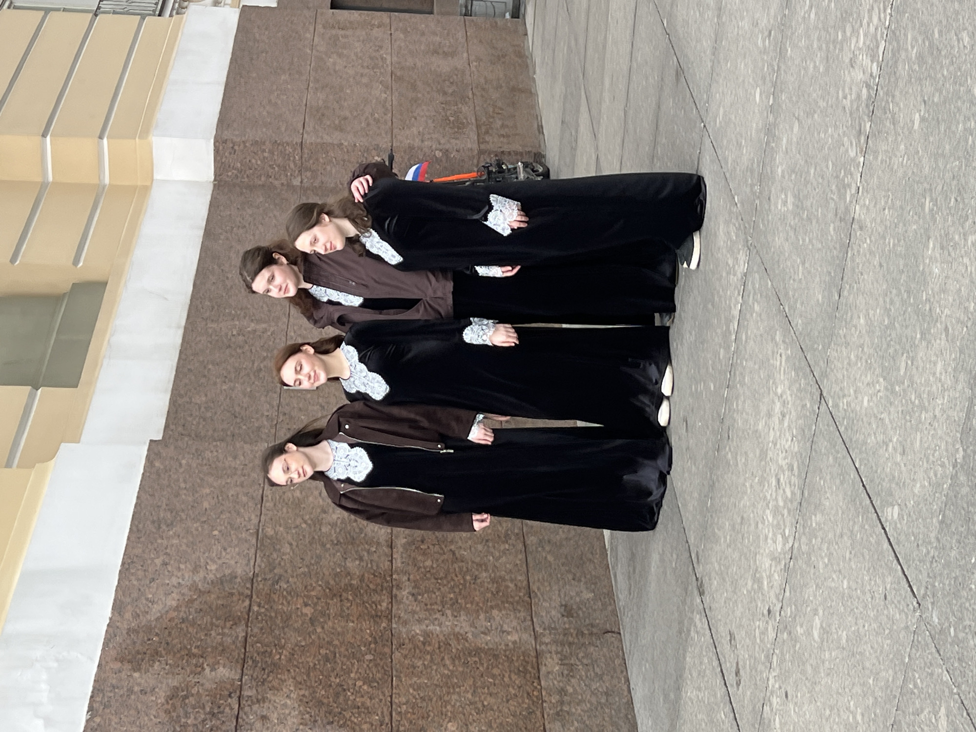 Four women stand in a row, posed against the backdrop of a building and a paved area in St. Petersburg, Russia. They are dressed in outfits that include dark, long-sleeved dresses with white lace collars and cuffs. Two of the women wear brown jackets over their dresses. The building features a base of rough-hewn brown material and a lighter-colored facade above. A parked vehicle with a Russian flag is visible behind the group. The paved area is gray and appears to be made of stone or concrete.