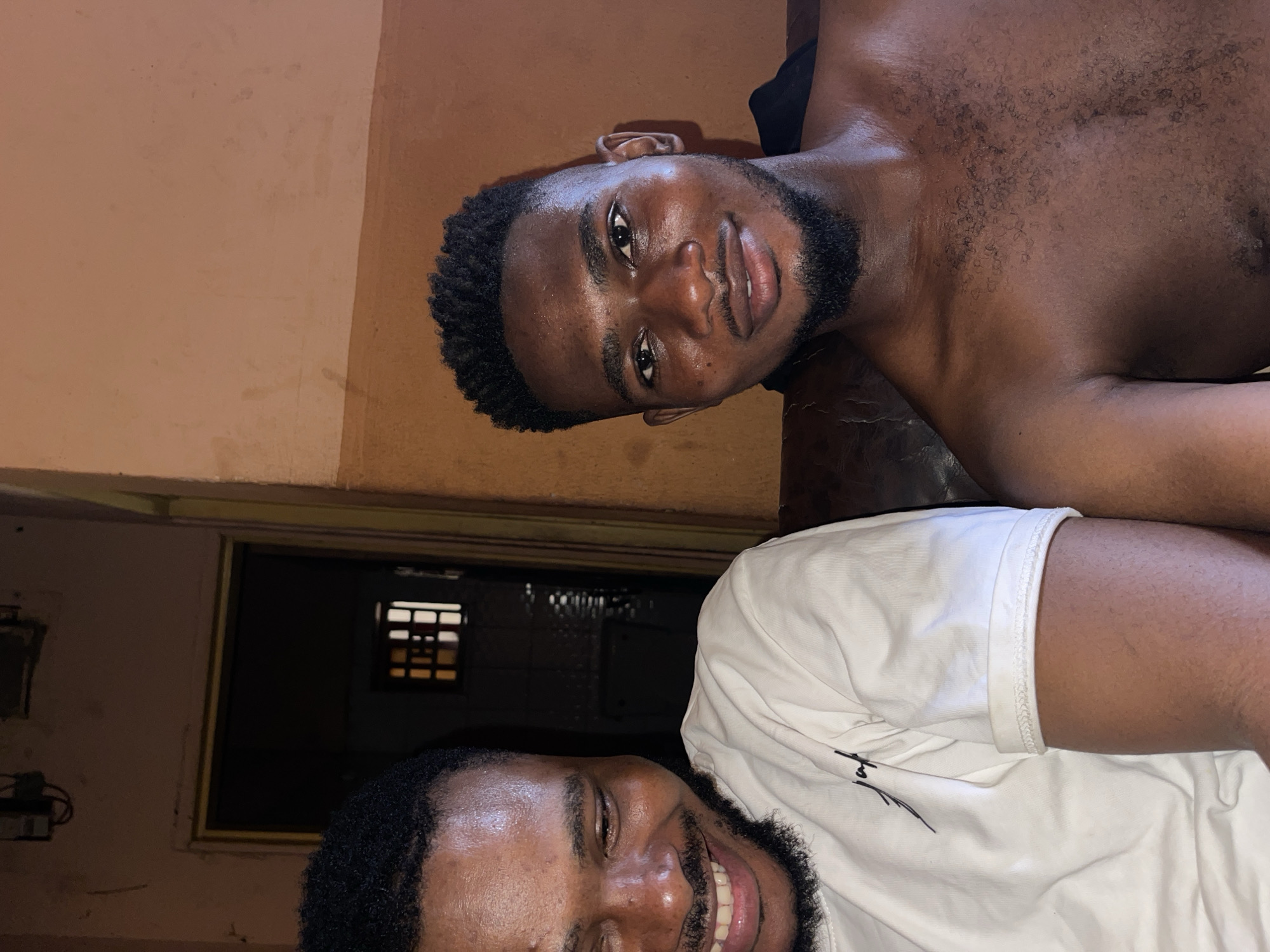 Two men are lying next to each other indoors. One man is shirtless with dark skin, a beard, and short curly hair. He is looking towards the viewer with a slight smile. The other man is wearing a white t-shirt and has a beard. He is also smiling. A wall is visible in the background, along with a door or opening with bars or a screen. The location appears to be indoors, in a residential area. The location in the city of Muchiya, Nigeria cannot be confirmed from the image alone.