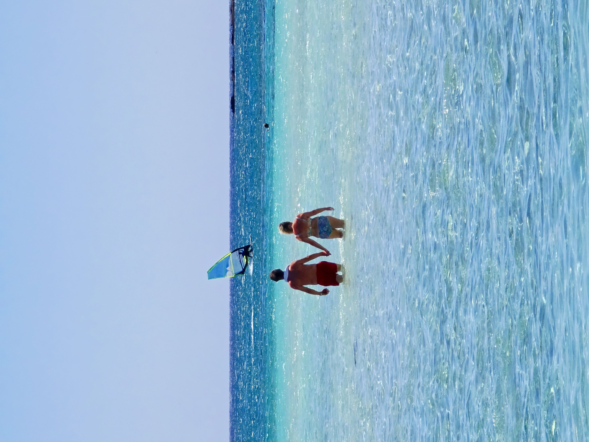 In the clear, turquoise waters of an unknown Greek location, a couple walks hand-in-hand, their bodies submerged up to their waists. The man wears red swim trunks, and the woman wears a patterned bikini. They are facing away from the camera, walking towards the deeper blue ocean. In the distance, a windsurfer is visible, their colorful sail a bright accent against the horizon. The water is calm, with gentle ripples catching the sunlight. The sky above is a clear, pale blue, with no clouds present.