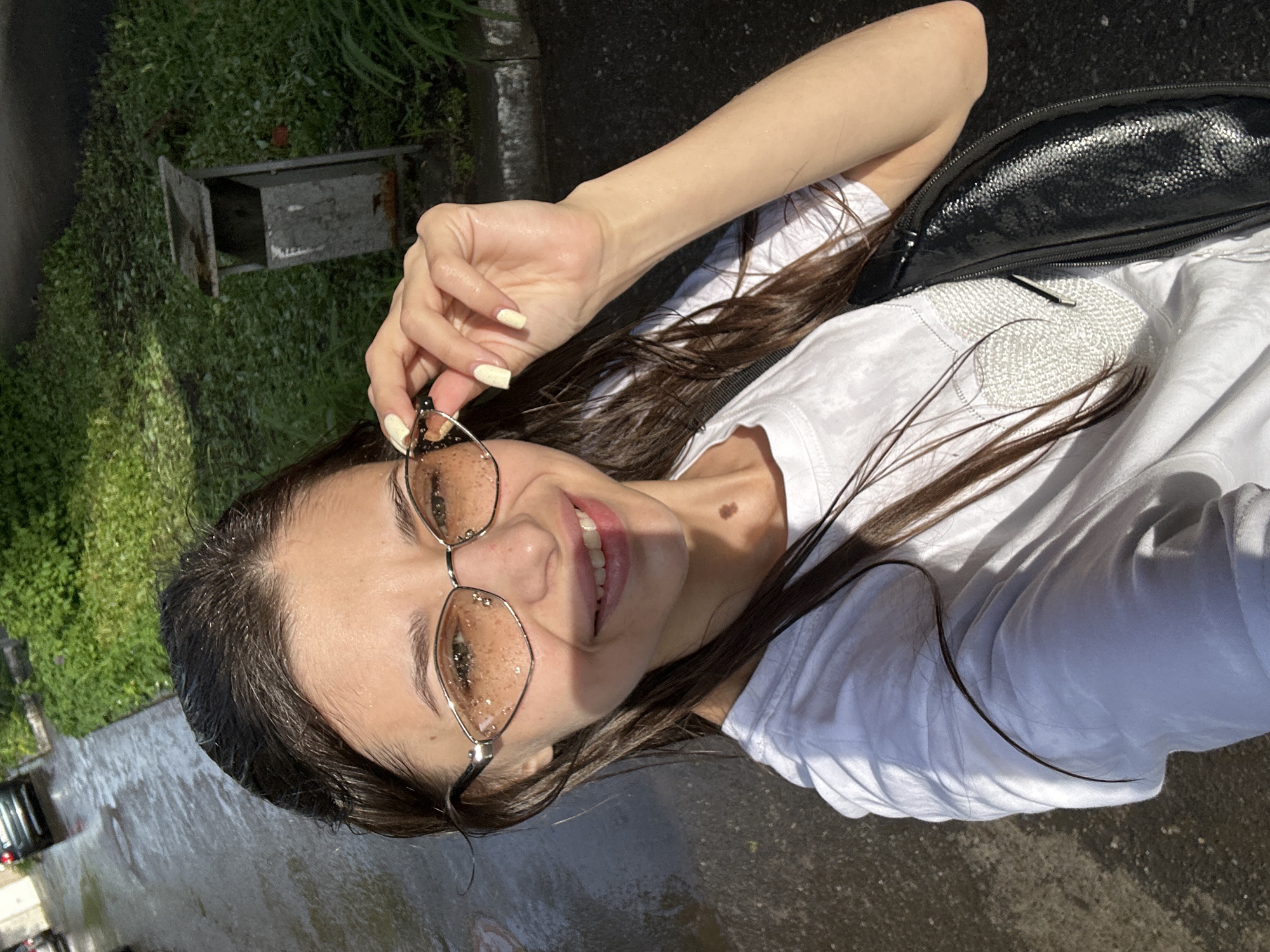 A young woman is photographed from a low angle, likely a selfie, outdoors in what appears to be a street environment. The background includes a building or structure to the left, along with greenery that could be trees or bushes. The ground appears to be pavement, possibly wet or reflective from recent rain.
The woman is wearing a white T-shirt and sunglasses with decorative elements inside the lenses. She's holding the sunglasses with her right hand. She has dark hair.
The image may have been taken near a specific city location, as the prompt suggests Izhevsk, Russia. However, visual markers to confirm a specific location are absent.