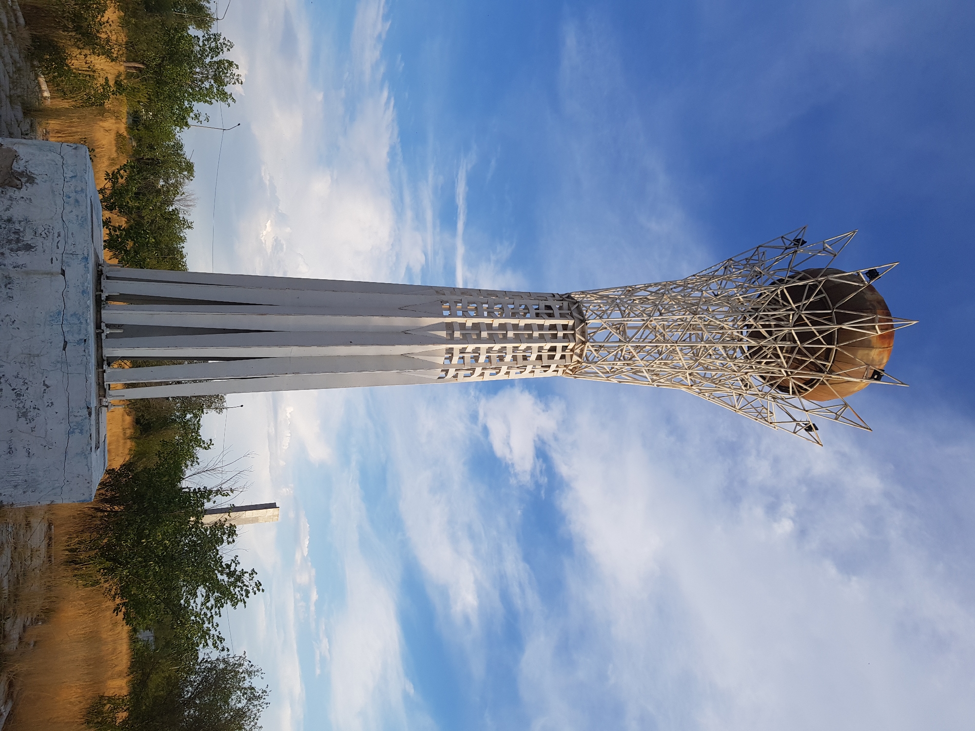 The image shows the Baiterek monument in Kurchatov, Kazakhstan, under a cloudy blue sky. The monument is a tall, white, metal structure with a spherical top made of a reddish-brown material. The base of the monument is a concrete pedestal. Surrounding the monument is dry, golden grass and several green trees. In the background, a tall, gray, rectangular structure is visible. The scene appears to be a public space.