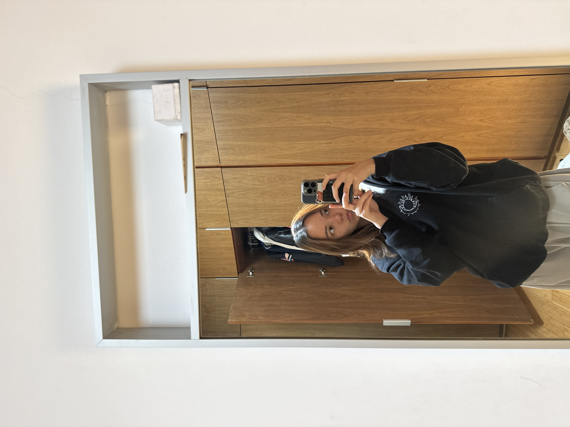 A woman is reflected in a framed mirror. She is holding a smartphone and taking a selfie. The mirror is mounted on a wall and reflects a wooden cabinet or wardrobe. The environment appears to be indoors, possibly a bedroom or dressing room. The location of San Bernardino Tlaxcalancingo, Mexico is not evident in the image.