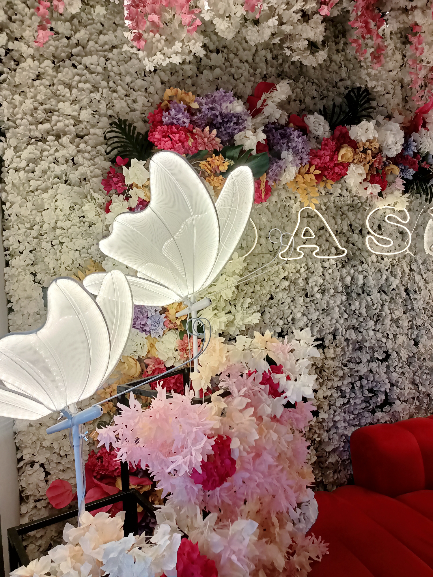 The image depicts a brightly decorated wall in Almaty, Kazakhstan, featuring a dense arrangement of artificial flowers in various colors including white, pink, red, purple, yellow, and orange. Two large, illuminated butterfly-shaped lamps are prominently displayed on the left side of the frame, casting a soft white glow. In the background, a neon sign spelling out "AS" is visible, also in white. To the right of the flower wall, a portion of a red, plush couch is visible, suggesting this is a seating area. The overall scene is vibrant and decorative, likely a photo backdrop or an interior design element.