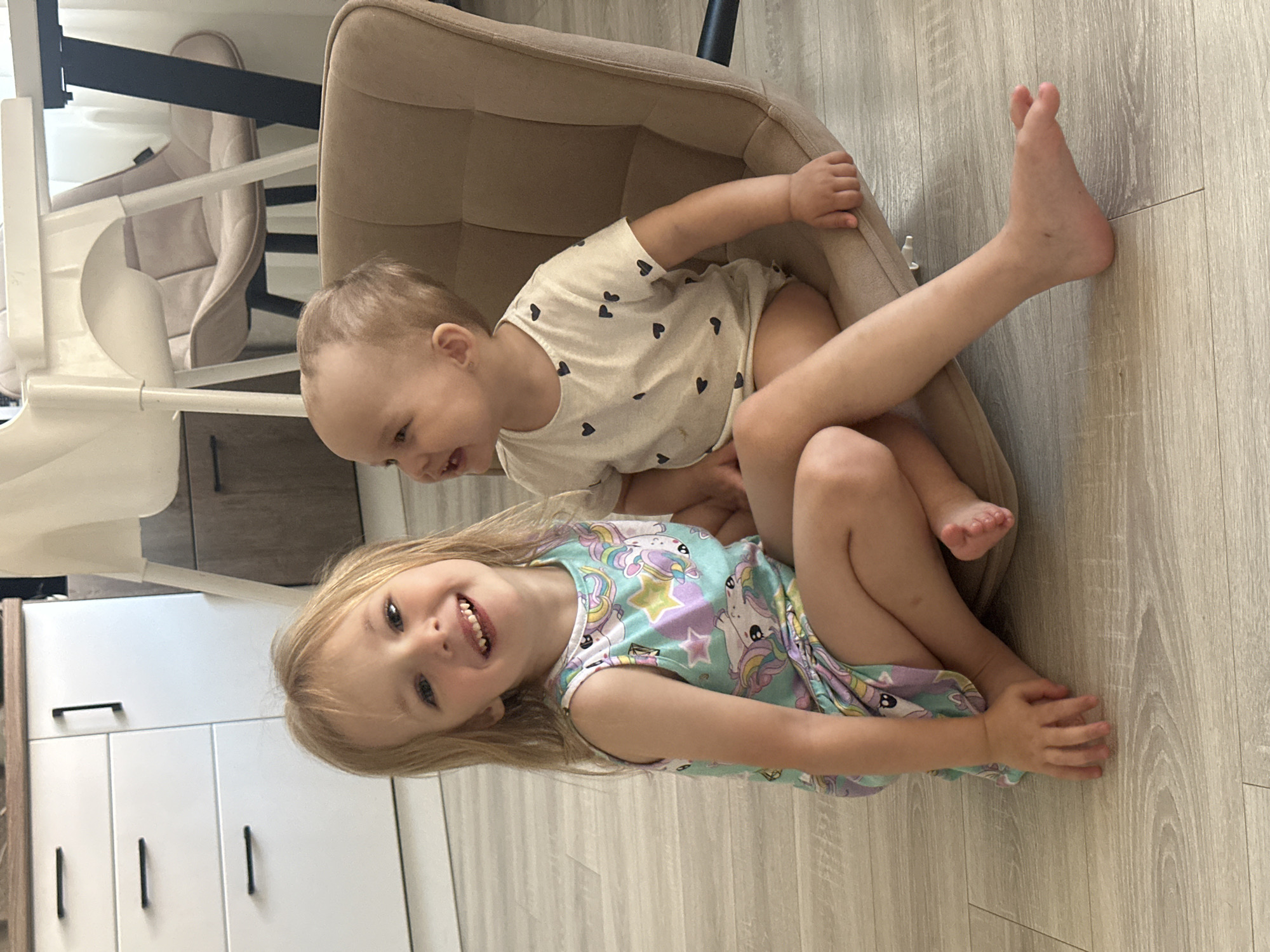 Two young children are seated in a beige armchair. The younger child, wearing a white shirt with black heart patterns, is situated within the chair. The older child, dressed in a unicorn-themed dress, is seated on top of the younger child, their feet extending outward. The setting appears to be inside a house, with a highchair and a white cabinet visible in the background. The location is implied to be in Chaikovsky, Russia.