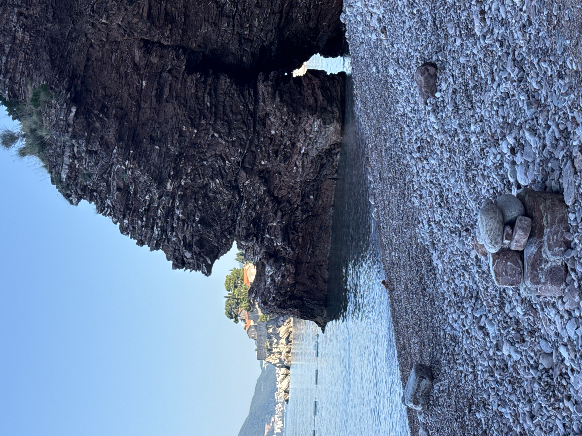This is a photograph of a rocky beach in Sveti Stefan, Montenegro. In the foreground, the beach is covered in small, smooth pebbles. A small cairn of stones is stacked on the right side of the foreground. In the middle ground, calm, clear blue water meets the shore. A large, dark rock formation with a natural archway dominates the right side of the frame, partially obscuring the view of a small island in the distance. This island, visible in the background, is covered with trees and appears to have buildings with red-tiled roofs. The island is connected to the mainland by a narrow causeway. The sky above is a clear, pale blue. The overall scene depicts a serene coastal landscape.