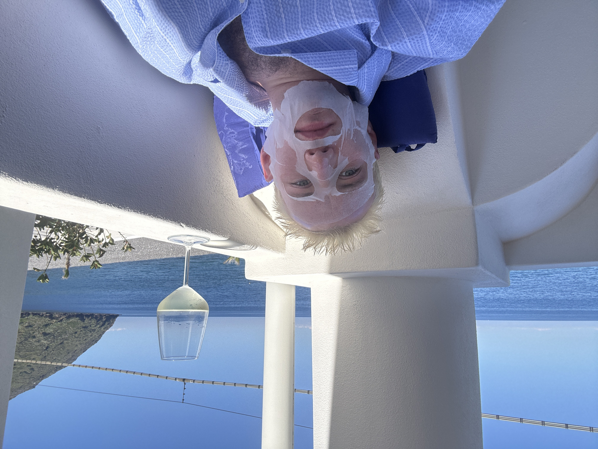 An inverted image shows a man relaxing on what appears to be a balcony or terrace overlooking a scenic coastal landscape in Lipari, Italy. The man, with blonde hair, is lying down, wearing a blue and white striped robe or shirt. He has a white sheet face mask covering his face, with cutouts revealing his open eyes and mouth. A blue pillow is visible beneath his head.
To his right, a stemmed glass, possibly a wine glass, filled with a light-colored liquid, rests on the balustrade. The background features a calm, vast expanse of blue sea meeting a clear blue sky. A large, green, mountainous island or landmass is visible in the distance to the left, rising from the water. White architectural structures, including a balustrade and a column, frame the view, suggesting a resort or hotel setting. Some green foliage is also visible near the balustrade. The bright lighting indicates it is daytime with clear, sunny weather, evoking a sense of leisure and self-care. There is no visible text in the image.