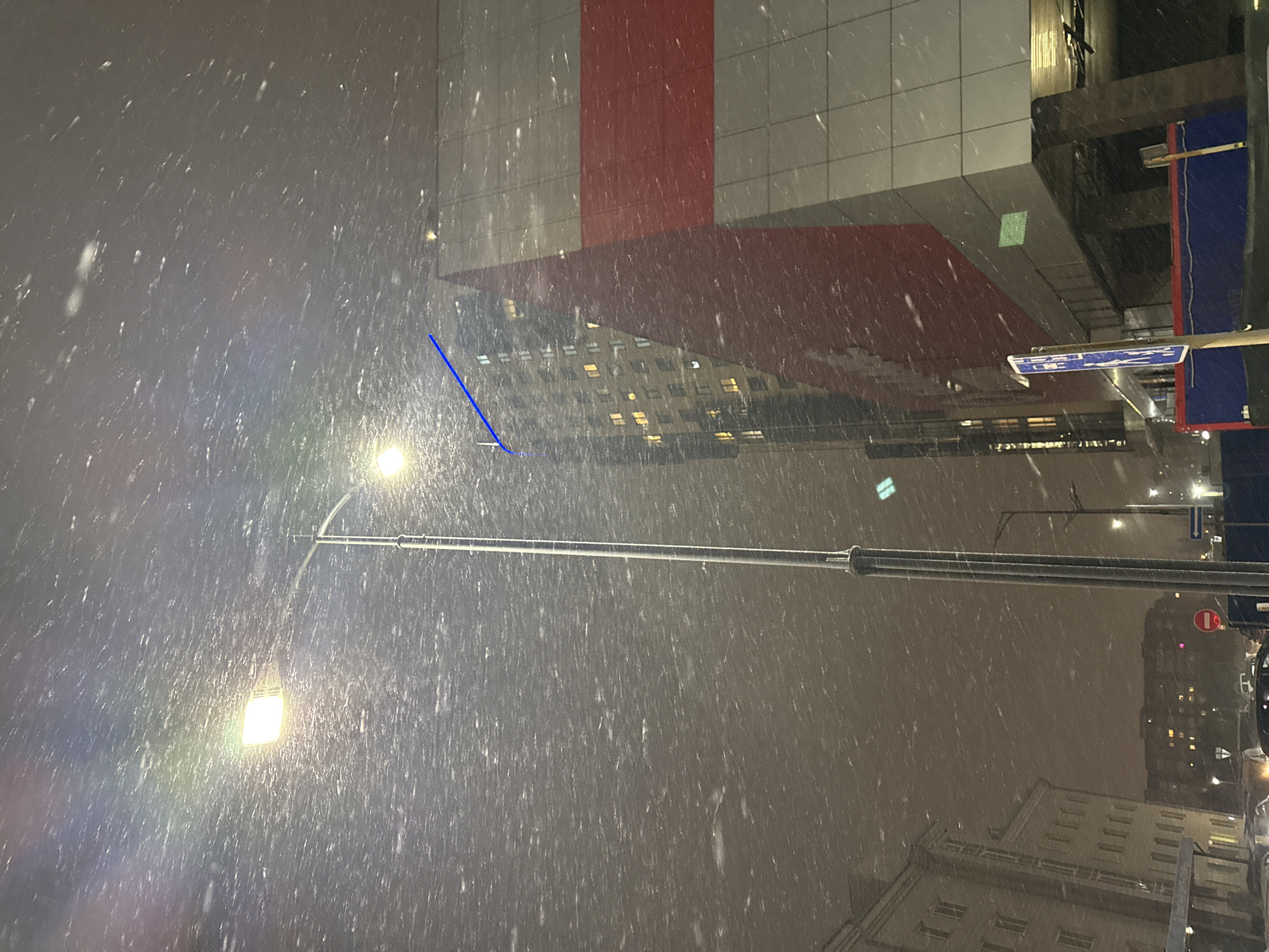 The image shows a snowy urban scene, possibly at night, in Krasnoyarsk, Russia. A street light is visible, illuminating falling snow. Buildings with lit windows are in the background. A modern building with a red and white facade is also present. A street sign is partially visible. The camera is likely positioned at a slight angle.