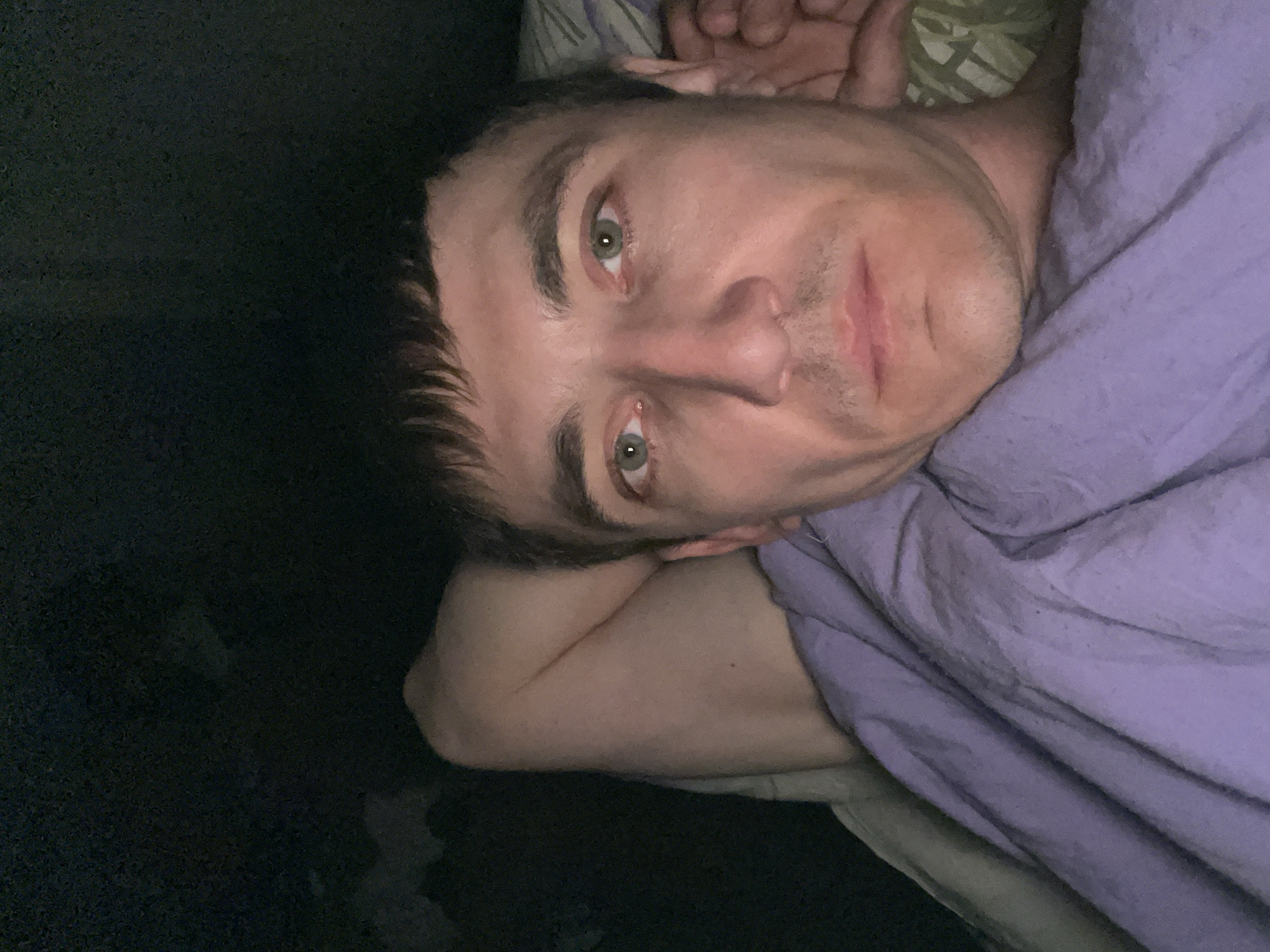 The image shows a close-up, overhead view of a man lying in bed. He has light skin, short dark hair, and green eyes. His left arm is bent and resting behind his head, and he is covered by a purple blanket. The background is dark, suggesting a dimly lit room. There are no visible elements in the image that would indicate a specific location in Laishevo, Russia.