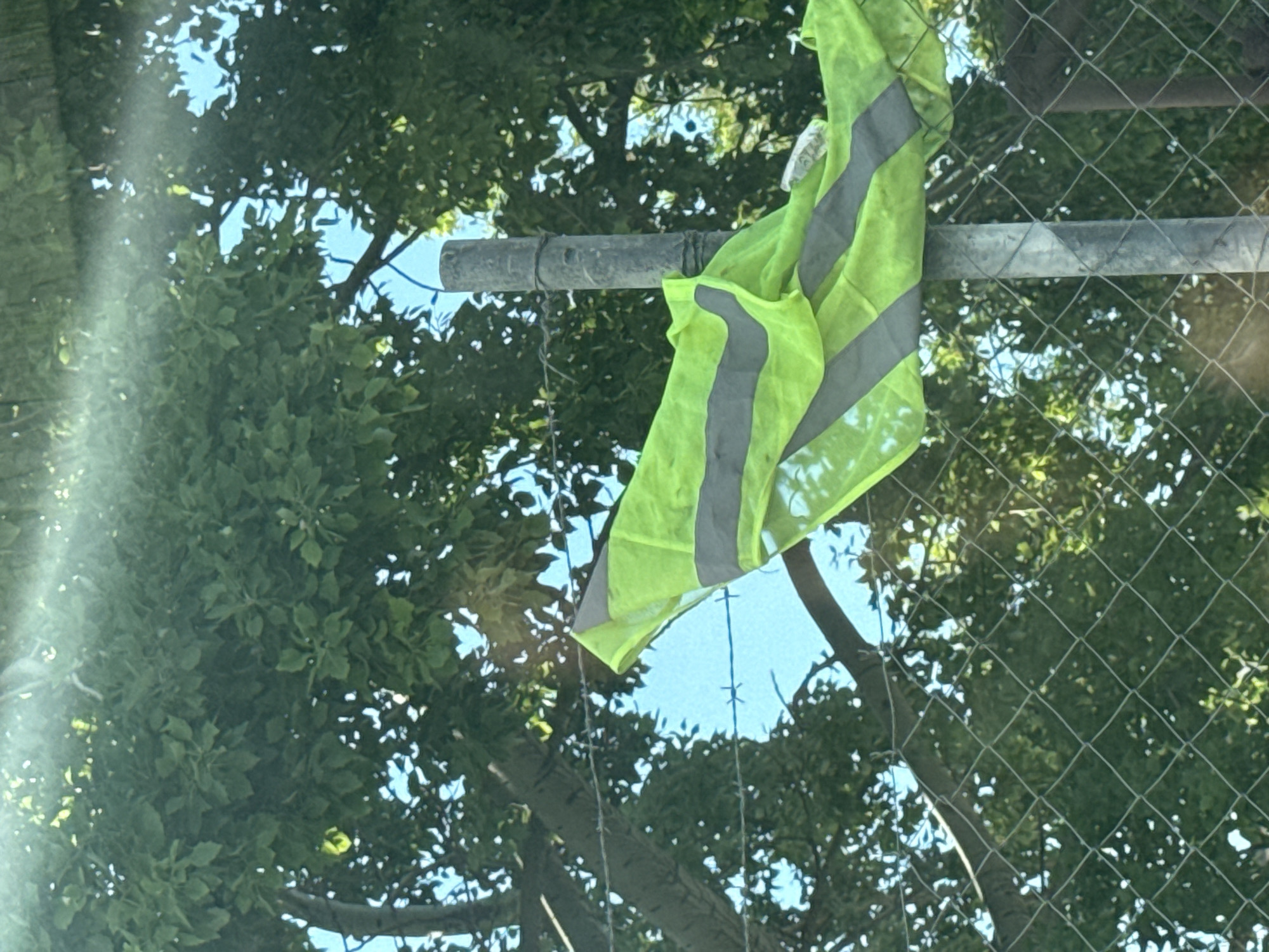 In Varna, Bulgaria, a fluorescent yellow safety vest with reflective grey stripes is draped over a barbed wire fence that is supported by a metal pole. The vest appears to be partially wet. In the background, lush green foliage from trees can be seen against a bright blue sky. A chain-link fence is also visible in the foreground, partially obscuring the background. The scene suggests an outdoor setting, possibly near a construction site or an area undergoing maintenance, given the presence of the safety vest.