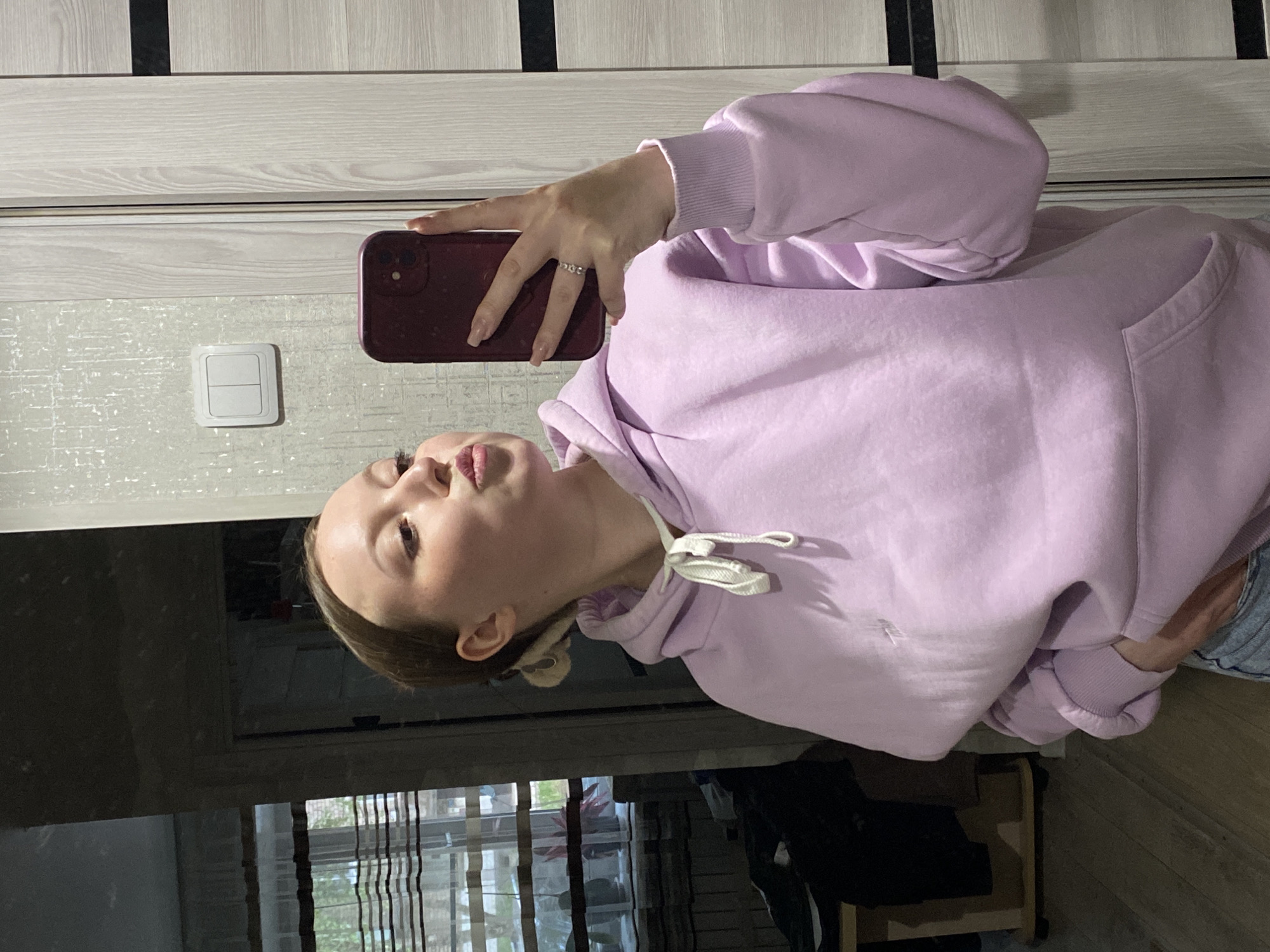 A young woman is taking a selfie indoors. She is wearing a light purple hoodie and holding a maroon-colored smartphone in her left hand. She has light skin and her hair appears to be pulled back. She is making a kissing face towards the camera. The background includes a light-colored wall and door with dark vertical accents, along with a light switch. A window with patterned curtains is visible. There is a chair in the bottom right corner of the image. There are no landmarks to confirm the location is Chaikovsky, Russia.