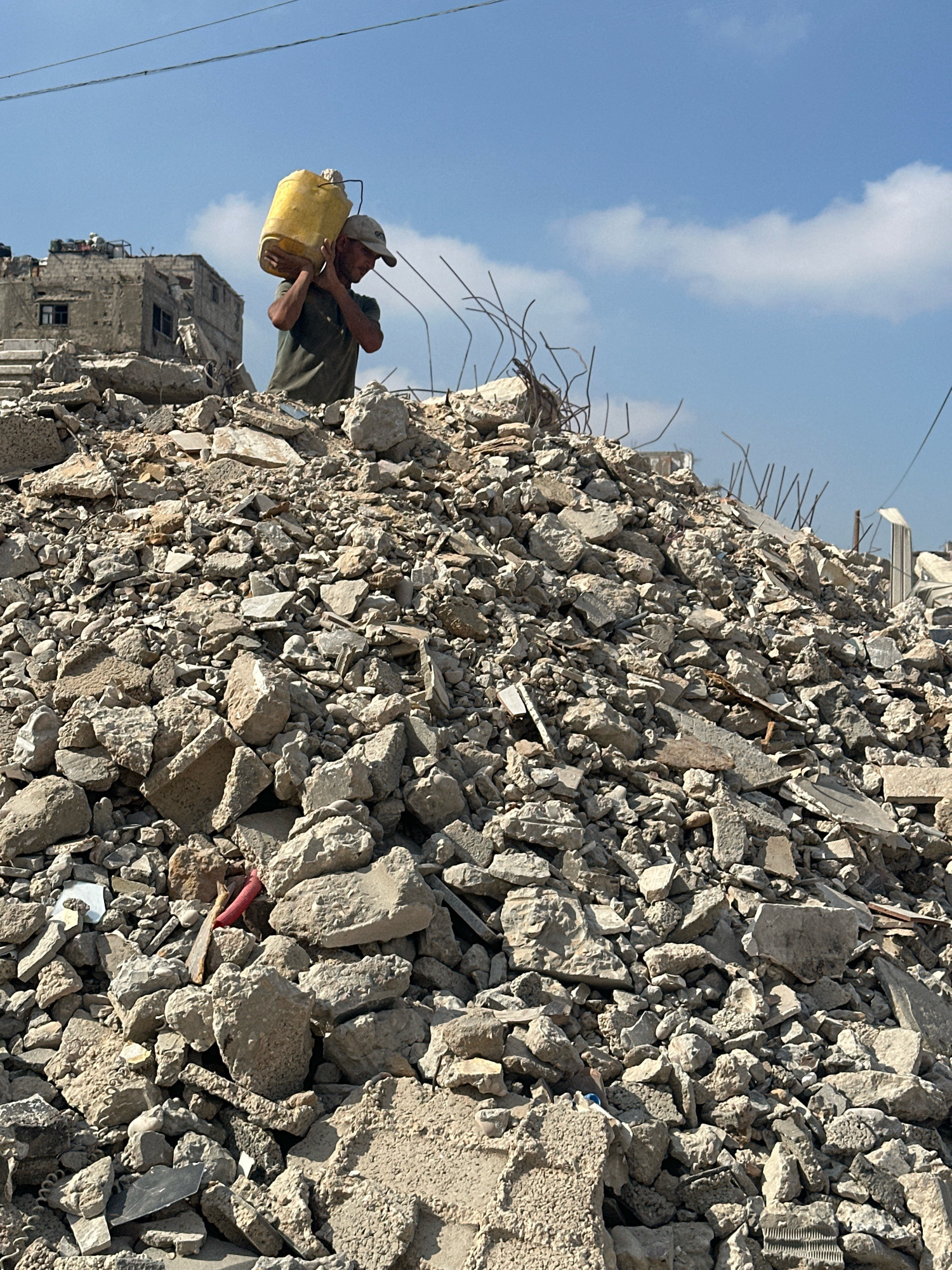 A man carries a yellow water jug over his shoulder as he walks across a pile of rubble. The rubble consists of broken concrete, rebar, and other debris. In the background, damaged buildings are visible under a blue sky with some clouds. The scene appears to be in the city of Gaza, Palestinian Territories, and suggests a post-conflict or disaster environment.