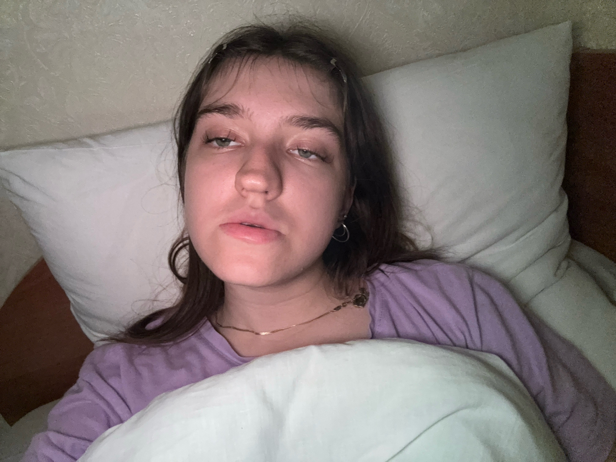 A young Caucasian female with medium brown hair is pictured lying in bed, looking towards the camera. Her eyes are partially closed, and her mouth is slightly open, giving her a sleepy or tired appearance. She is wearing a purple long-sleeved shirt and has a thin gold necklace. A white pillow is positioned behind her head, and another light-colored blanket or sheet is pulled up to her chest. The background consists of a textured wall and the edge of a brown headboard. The lighting is dim, suggesting it may be nighttime or early morning. The image was taken indoors. The location is not specified within the image itself, and therefore, cannot be determined to be Zavolzhye, Russia.