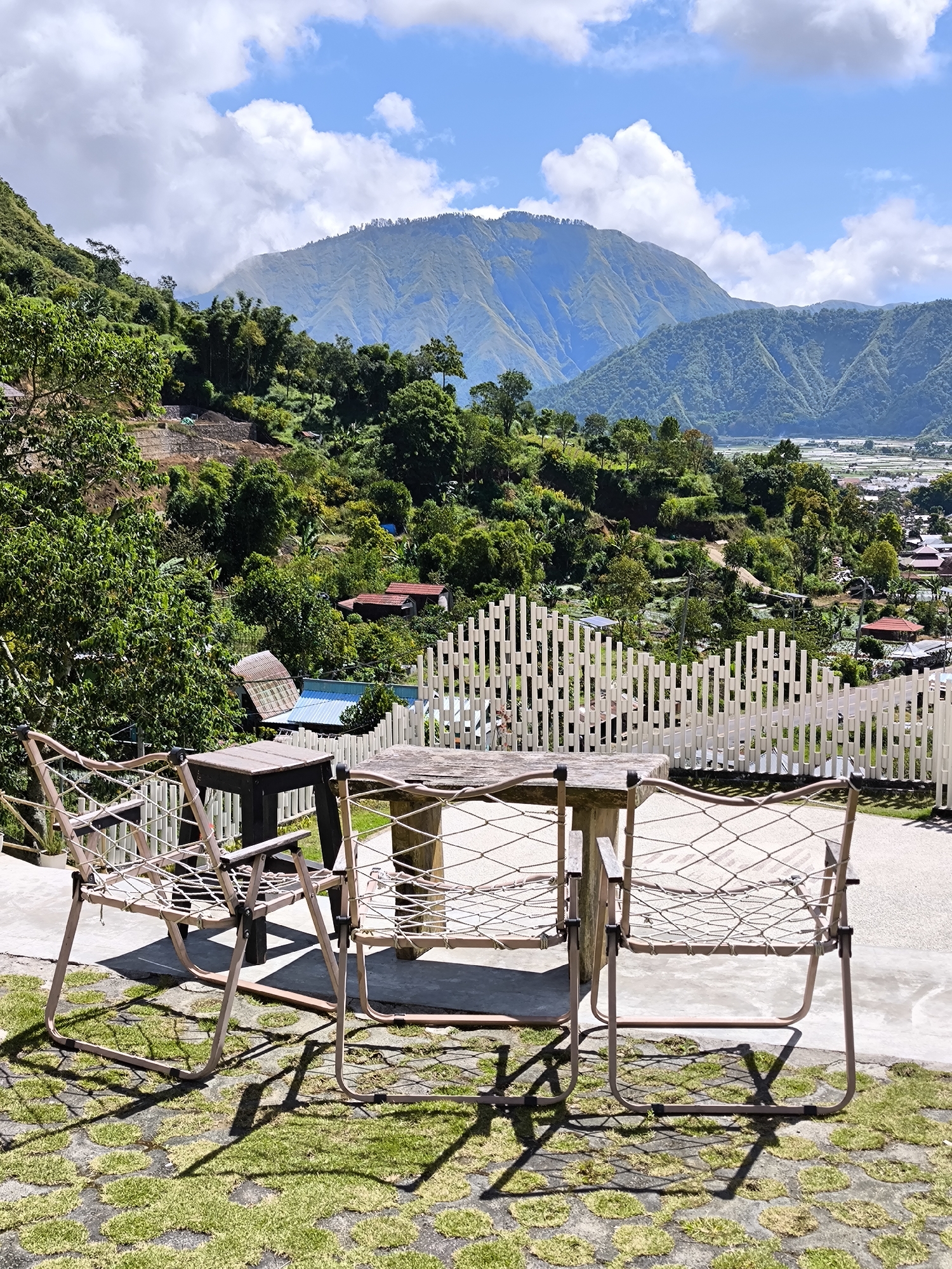 The image shows an outdoor seating area in Sembalun Bumbung, Indonesia. The seating area consists of a weathered wooden table and two matching chairs with a rope-like weave design for the seats and backs. There is a small black side table next to one of the chairs. The seating is placed on a stone-paved surface interspersed with patches of grass. A white, modern-style fence with vertical slats is visible in the background. Beyond the fence, there is a view of a village with scattered houses, lush green trees, and terraced hillsides. In the distance, a large mountain range is visible under a blue sky with scattered white clouds.