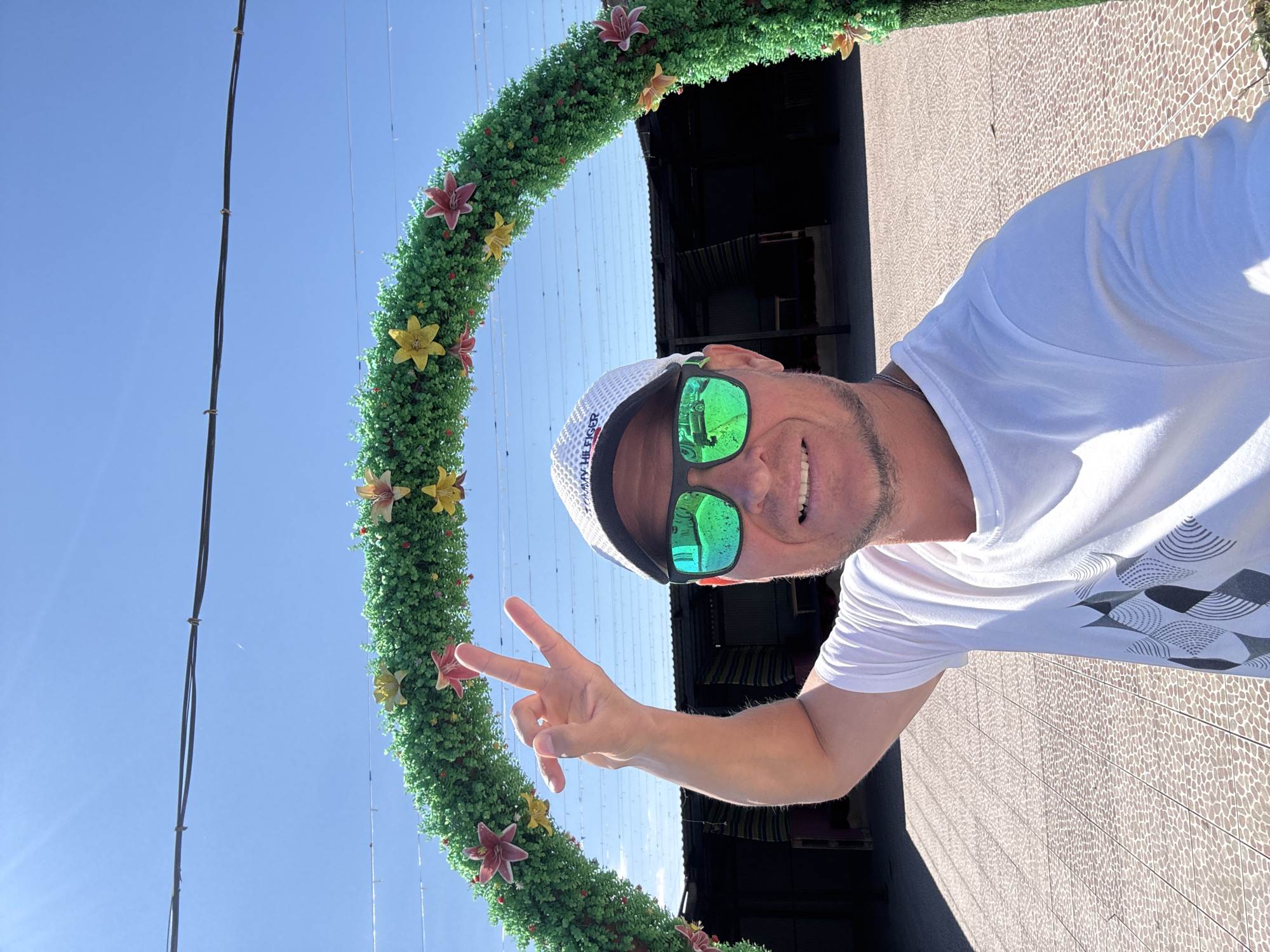 A man, wearing a white t-shirt, a white cap with a Tommy Hilfiger logo, and green mirrored sunglasses, poses for a selfie. He is standing in front of a green, artificial floral arch decorated with red and yellow flowers. Behind the arch, there are dark structures, possibly market stalls or covered areas, with striped awnings. The ground is paved with small, light-colored tiles arranged in a mosaic pattern. The sky is clear and blue, with some visible power lines overhead. The man is smiling and making a gesture with his right hand, with his thumb and index finger extended and his middle and ring fingers bent down. The overall setting appears to be an outdoor marketplace or recreational area in the city of Urdzhar, Kazakhstan.