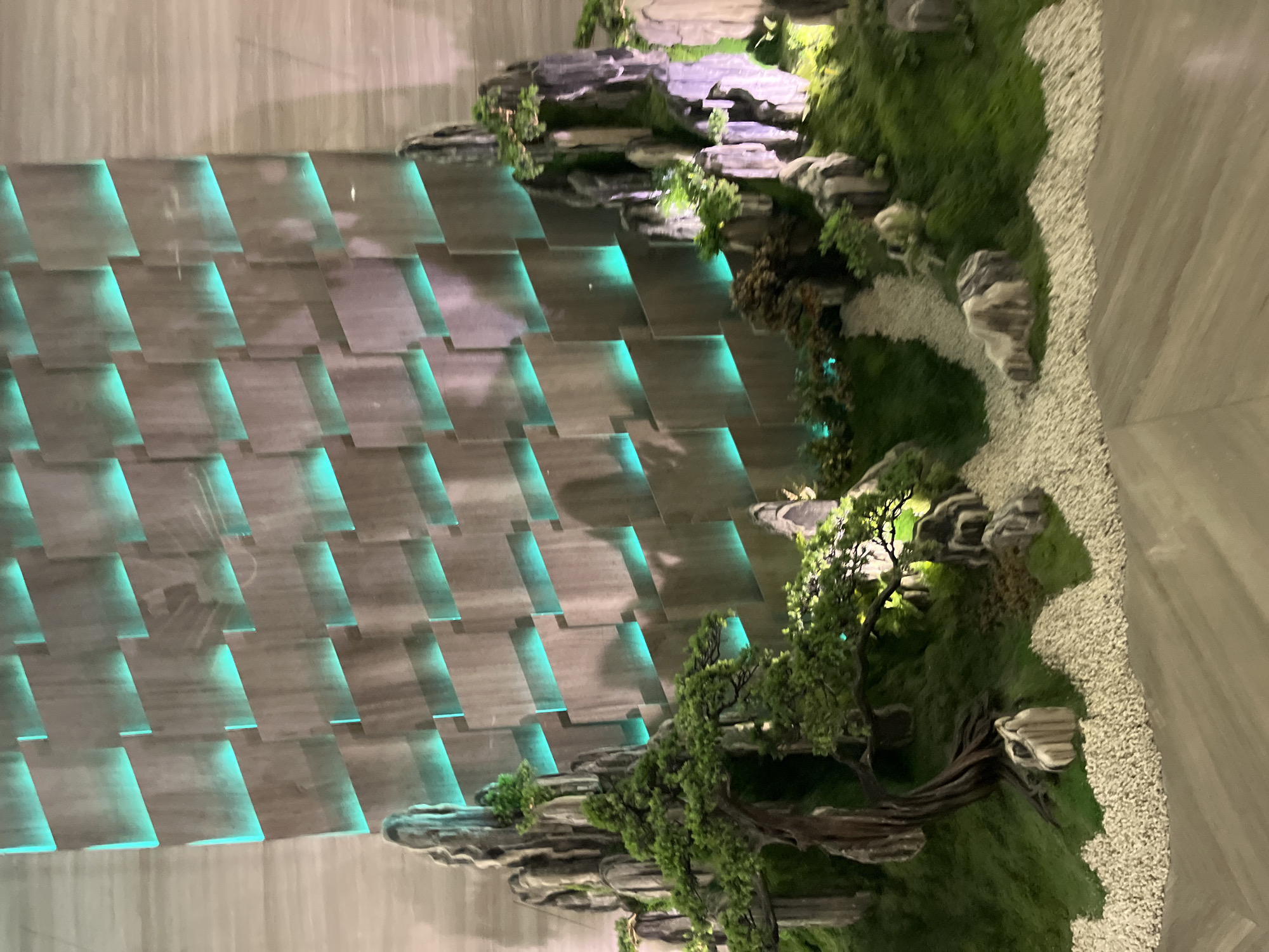 This media file showcases a meticulously crafted miniature landscape, resembling a Zen garden or a rock garden, positioned against a unique, illuminated wall. The wall is composed of staggered, rectangular panels that create a textured, layered effect. Behind these panels, soft, teal-colored lights emanate, casting an ethereal glow that highlights the geometric patterns of the wall.
The miniature landscape itself features artificial rocks of various sizes and textures, some covered in lush green moss and vegetation. Small, intricately detailed trees with dark, twisted branches and dense green foliage are strategically placed among the rocks. White gravel forms a winding path or area within the landscape, adding to the serene aesthetic. The overall impression is one of tranquility and natural beauty, artfully presented indoors.
The scene is likely set in a contemporary indoor space, perhaps a lobby, a spa, or a high-end restaurant in Nha Trang Ward, Vietnam, given the provided location context. The lighting, with its cool teal tones, suggests an evening or nighttime setting, or simply an intentional ambiance to enhance the visual experience of the display. There are no people visible in the media file, and no visible text. The focus is entirely on the artistic arrangement of the rock garden and the striking backdrop.