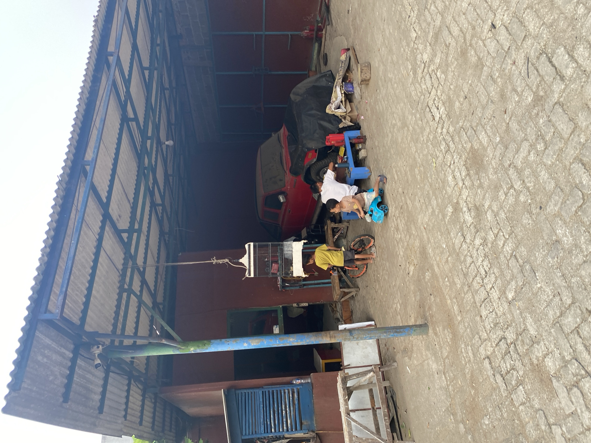 This image depicts an outdoor scene under a corrugated metal roof supported by a metal frame, likely a workshop or garage, in Tangerang, Indonesia. In the foreground, a paved area with interlocking stones shows signs of wear and tear. A young boy in a yellow t-shirt stands next to a small red bicycle, facing towards the right. Further into the scene, a second young boy is seated on a blue toy car, pushing himself forward. To his right, another individual, likely an older boy or young man wearing a white t-shirt, is working on a red truck partially covered by a black tarp. Various objects are scattered around the workshop, including tools, containers, and a birdcage hanging from a rope. The background consists of a red building with blue window frames. The overall impression is a functional, somewhat cluttered, outdoor workspace.