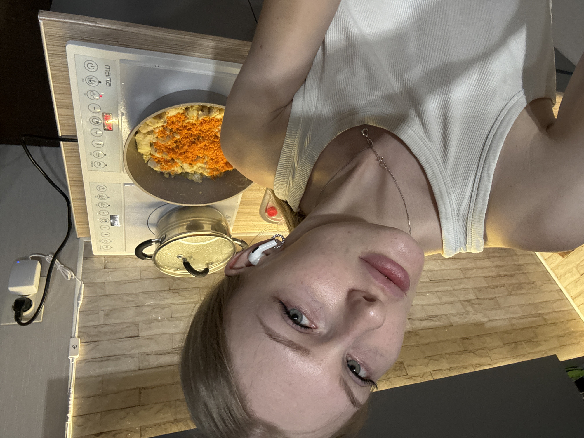 A woman is positioned in a kitchen environment. She is wearing a white tank top. In the background, a stove is visible with a pan on top containing food with orange toppings. Next to the pan, there is a pot covered with a lid. A power outlet and switch are seen on the kitchen wall. The location may be Perm, Russia, as suggested.