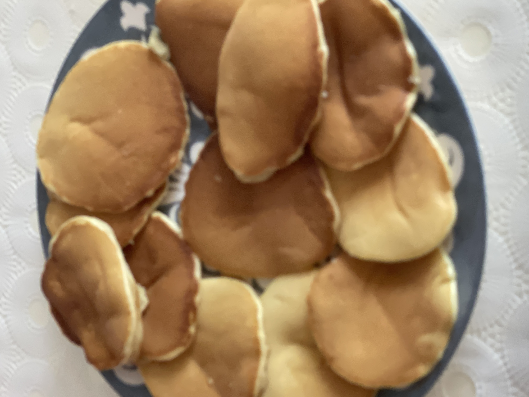 A plate of pancakes is shown from a top-down view. The pancakes are golden brown with some lighter golden brown edges. They are piled on top of each other on a dark blue plate with a white pattern. The plate is on a white, textured surface, likely a tablecloth. The image appears to be taken in a home setting in Sumgayit, Azerbaijan.