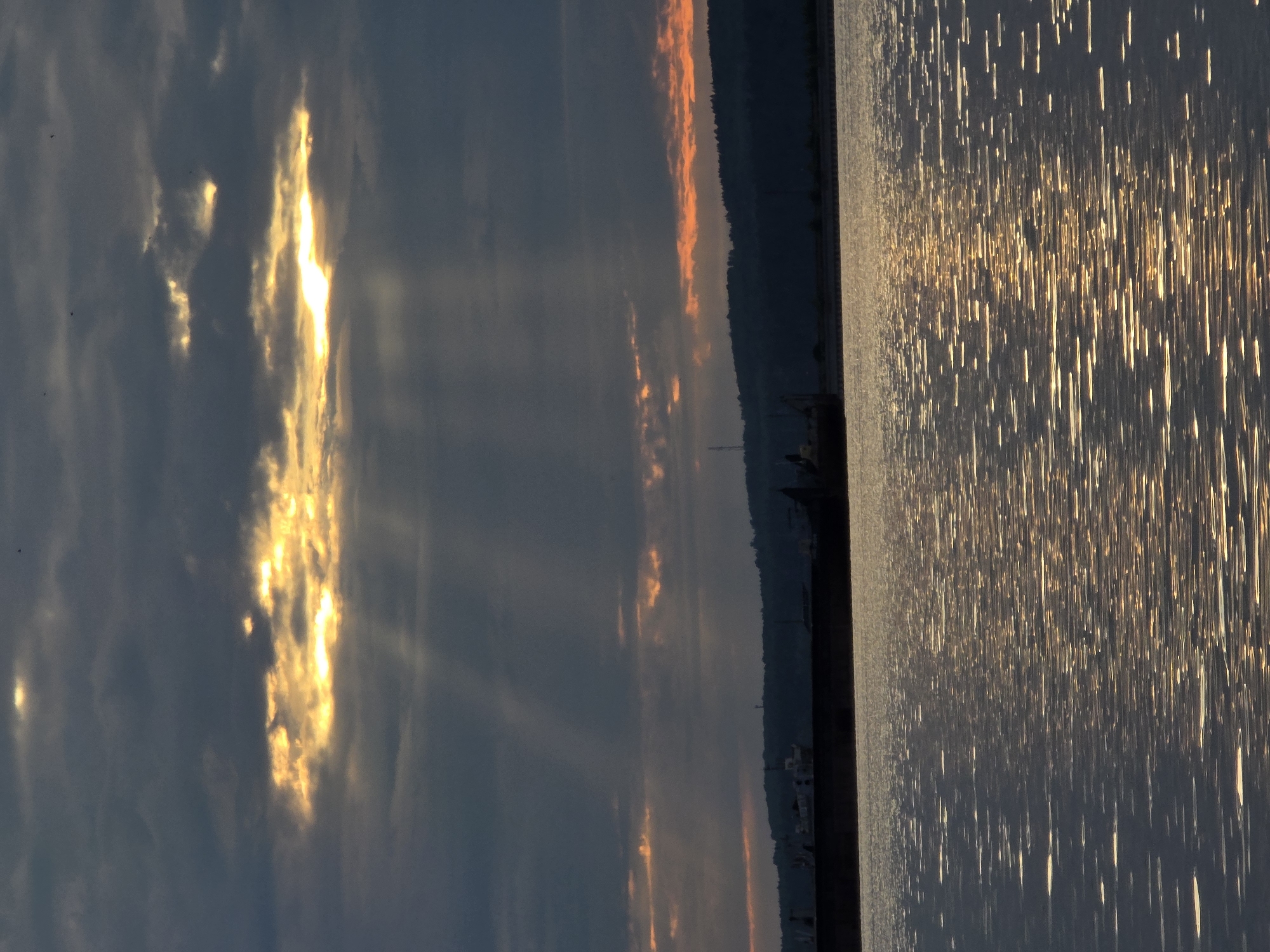 The image depicts a serene, overcast sky over the Kama River in Chaikovsky, Russia. Sunbeams pierce through the clouds, creating a dramatic shaft of light that illuminates the water's surface with shimmering reflections. In the distance, the silhouette of a forested hill forms the horizon, with faint hints of man-made structures visible along its crest. A large barge or ship is partially submerged in the foreground, its dark hull contrasting with the glinting water. The overall atmosphere is one of peaceful twilight, with the muted colors of the sky and water creating a somber yet beautiful natural landscape.