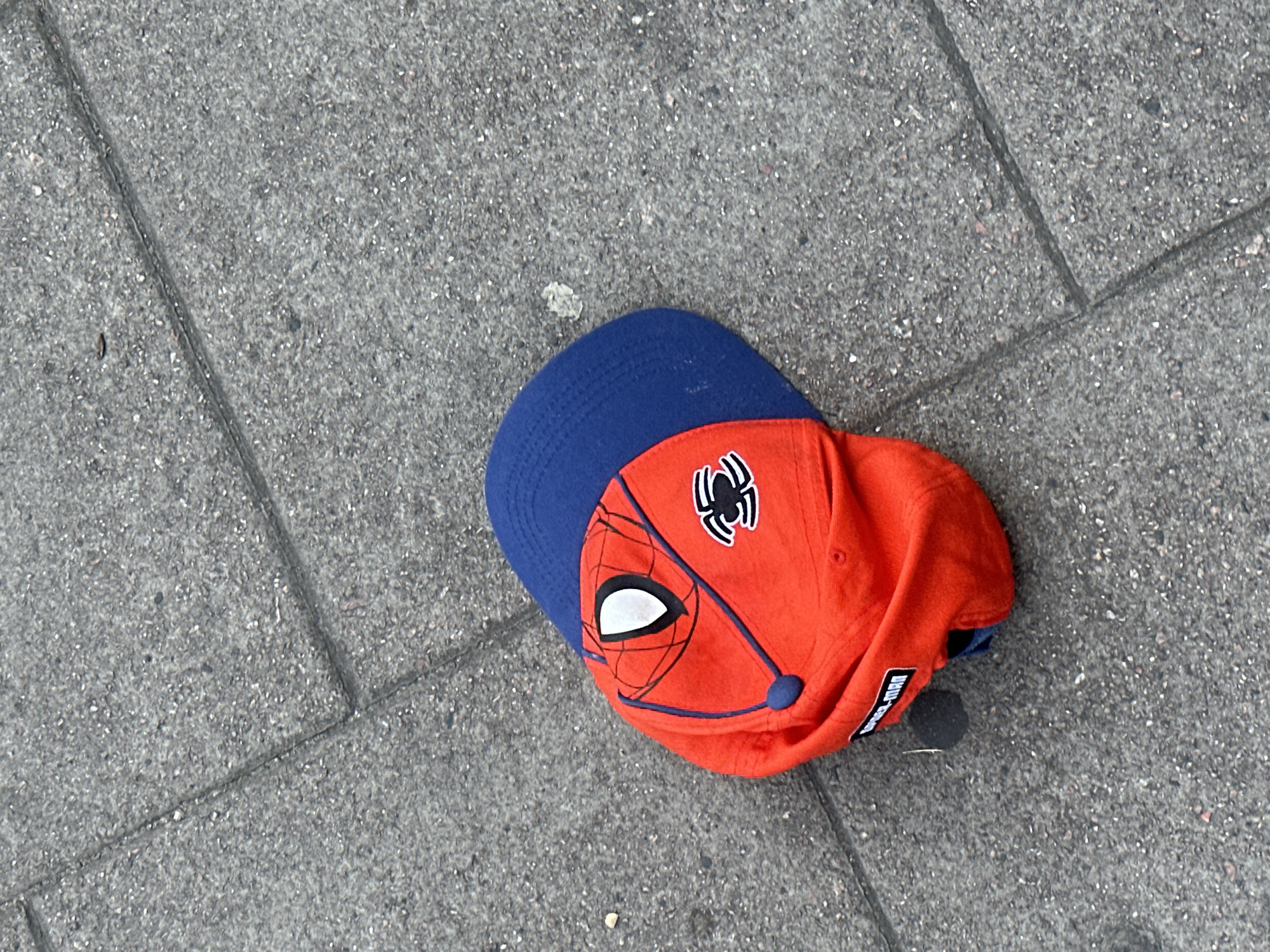 A Spiderman-themed baseball cap lies on a paved surface. The cap has a blue brim and a red crown emblazoned with a Spiderman mask design. The paved surface is grey and patterned with rectangular blocks. The location cannot be determined from visual information.