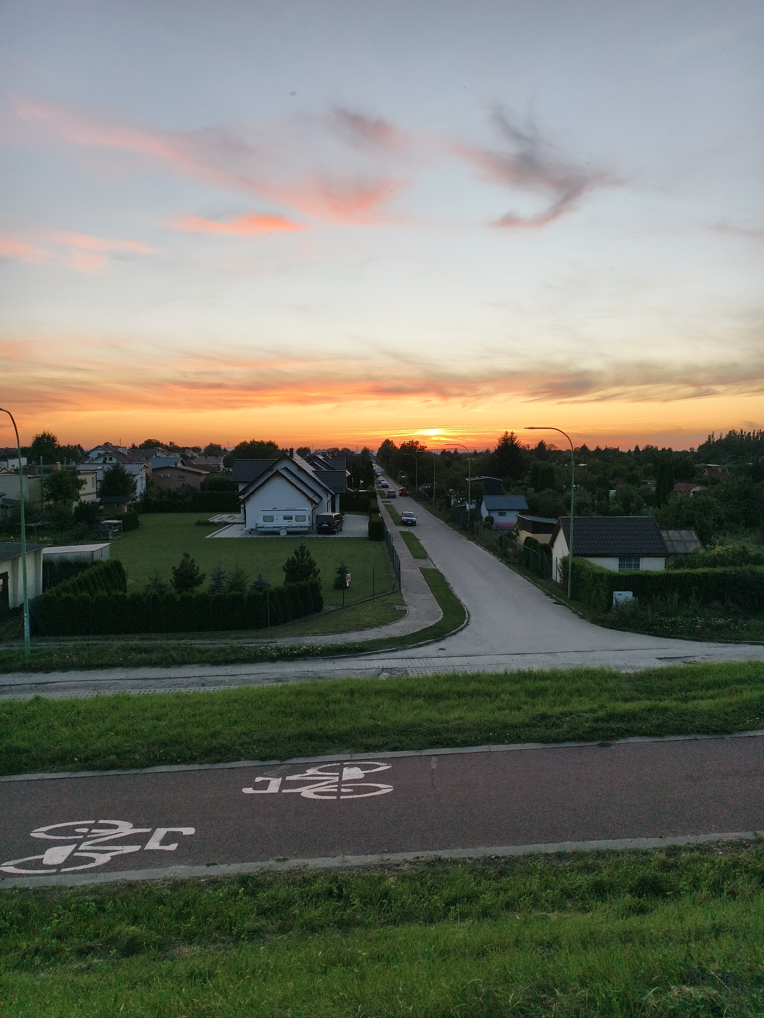 An eye-level, high-angle, full shot captures a street in Tczew, Poland, during a sunset. The sky is a gradient of colors, with soft pink and orange clouds scattered across a pale blue expanse. The sun is visible on the horizon, casting a warm glow over the scene. A street lined with houses and trees stretches into the distance, with a few cars parked along its side. In the foreground, a bicycle lane is marked with white symbols on a dark asphalt surface. The lane is bordered by a grassy slope.