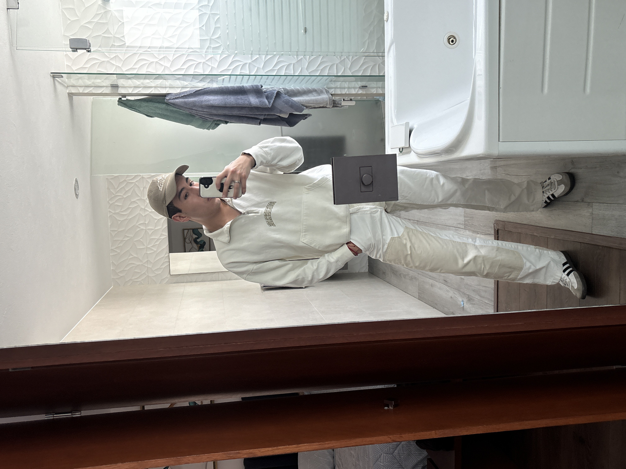 A young man is depicted in a full-body mirror selfie, captured in what appears to be a modern bathroom or dressing area located in San Bernardino Tlaxcalancingo, Mexico. He stands facing the mirror, holding a smartphone with a distinctive black and white cow-print case to take the photograph, with his left hand casually in his pocket and a ring visible on his ring finger.
He is dressed in a casual, monochromatic ensemble featuring a light-colored, possibly off-white or beige, long-sleeved top with a half-zip closure. Visible text on the front of the top appears to read "PREMIERE." He pairs this with matching light-colored trousers and a beige baseball cap, which has faint embroidered text, possibly "ADVENTURE." His footwear consists of white sneakers with black stripes, resembling Adidas Sambas.
The reflected environment is brightly lit and features contemporary design elements. The background includes white textured wall tiles with an organic or leafy pattern, a glass shower partition, and a towel rack holding two folded towels in grey and light green/blue hues. A white bathtub or jacuzzi is visible to the right. The floor is covered in light-toned, wood-effect tiled flooring. A dark, rectangular control panel with a circular button is reflected on the man's midsection. The mirror is framed by dark brown wood, and below it, an open wooden cabinet or vanity door reveals internal shelving.