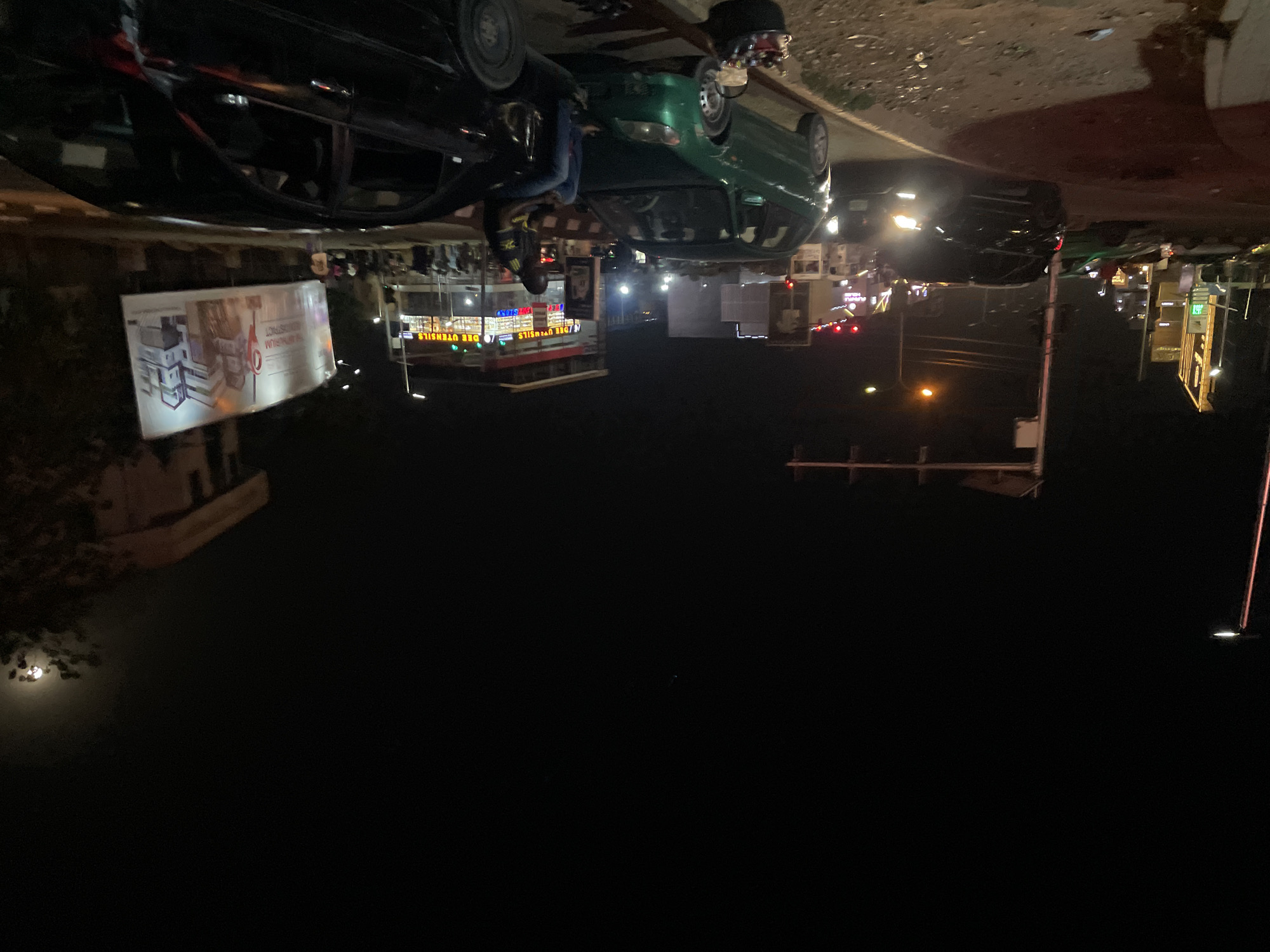 The image captures a vibrant and unusual night scene on a commercial street in Abuja, Nigeria. The central focus is an elaborate display featuring two full-sized cars, a black sedan and a green sedan, both conspicuously suspended upside down from an unseen overhead structure. Their wheels point skyward. A person, wearing a black and yellow shirt and blue pants, is positioned near the black inverted car, also appearing to be upside down, possibly interacting with the display or part of it.
Beneath and around this unique installation, the street is bustling with night-time activity. Numerous brightly lit stalls or small shops line the roadside, showcasing goods. Further down the street, another black car is visible, moving in its normal orientation with headlights on, indicating active traffic. The background shows faint outlines of distant buildings and structures against a dark night sky, where hints of a bright light source, possibly the moon, can be seen.
The scene is dramatically illuminated by a myriad of artificial lights, including bright shop lights, streetlights, and car headlights, creating stark contrasts between brightly lit areas and deep shadows. The unusual display of inverted cars suggests an art installation, a promotional stunt, or an educational exhibit, creating a surreal focal point within the busy urban environment.
Visible text on a large, illuminated banner hanging near the black suspended car reads "TRAUMA DISTRICT" (displayed upside down on the banner). Another shop sign beneath the display clearly shows "DRE UTENSILS" (also appearing upside down on the sign).