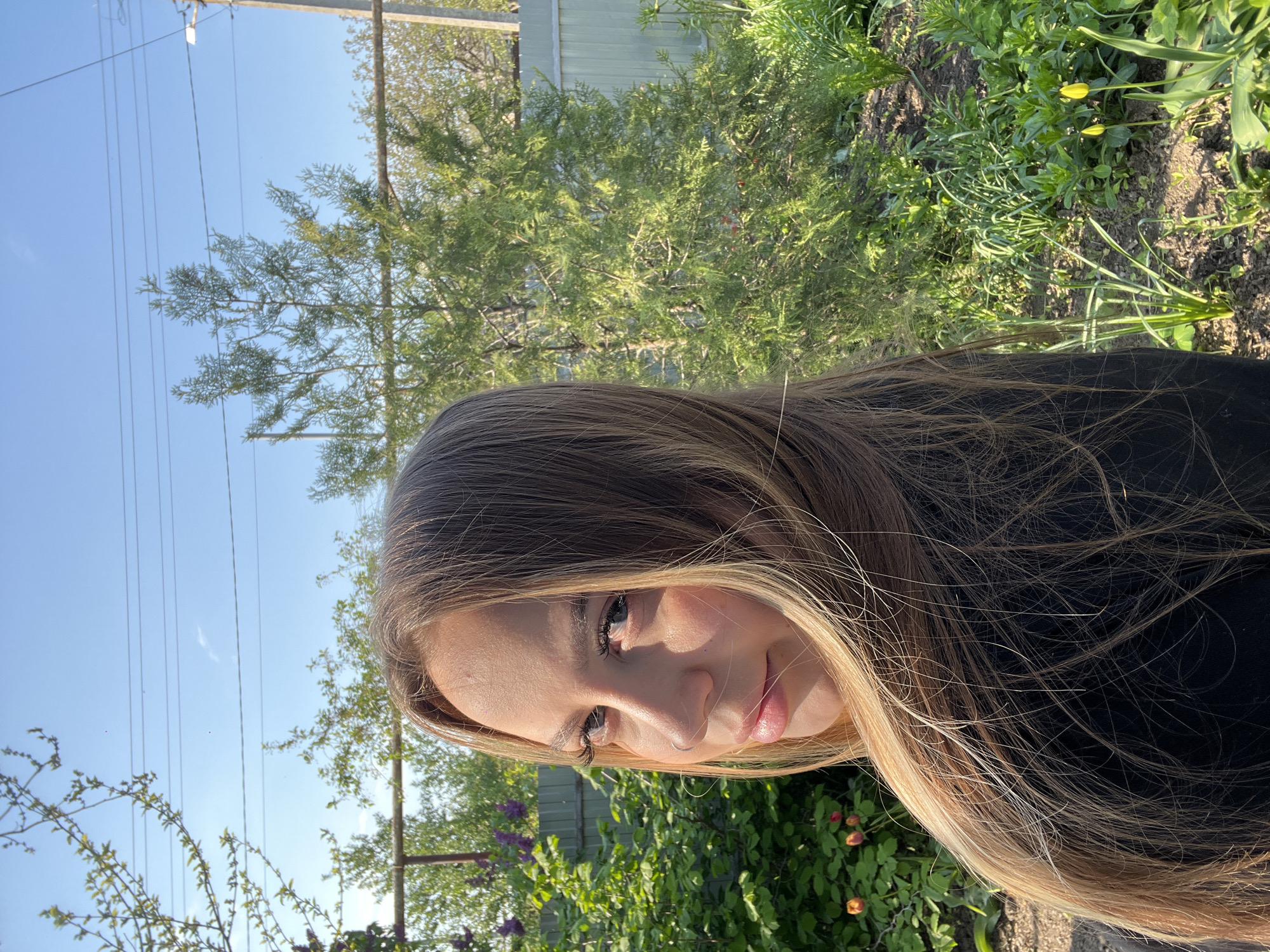 A young woman with long, blonde-streaked brown hair is positioned in the foreground, partially obscuring the scene. The location appears to be an outdoor setting, possibly a garden or yard. In the midground, there is a green, leafy area with a mix of plants and some visible flowers, including purple and red blossoms. A wooden fence or structure is partially visible behind the greenery. The background shows a clear, blue sky with telephone poles and wires spanning across. This setting is in Novopokrovskaya, Russia.