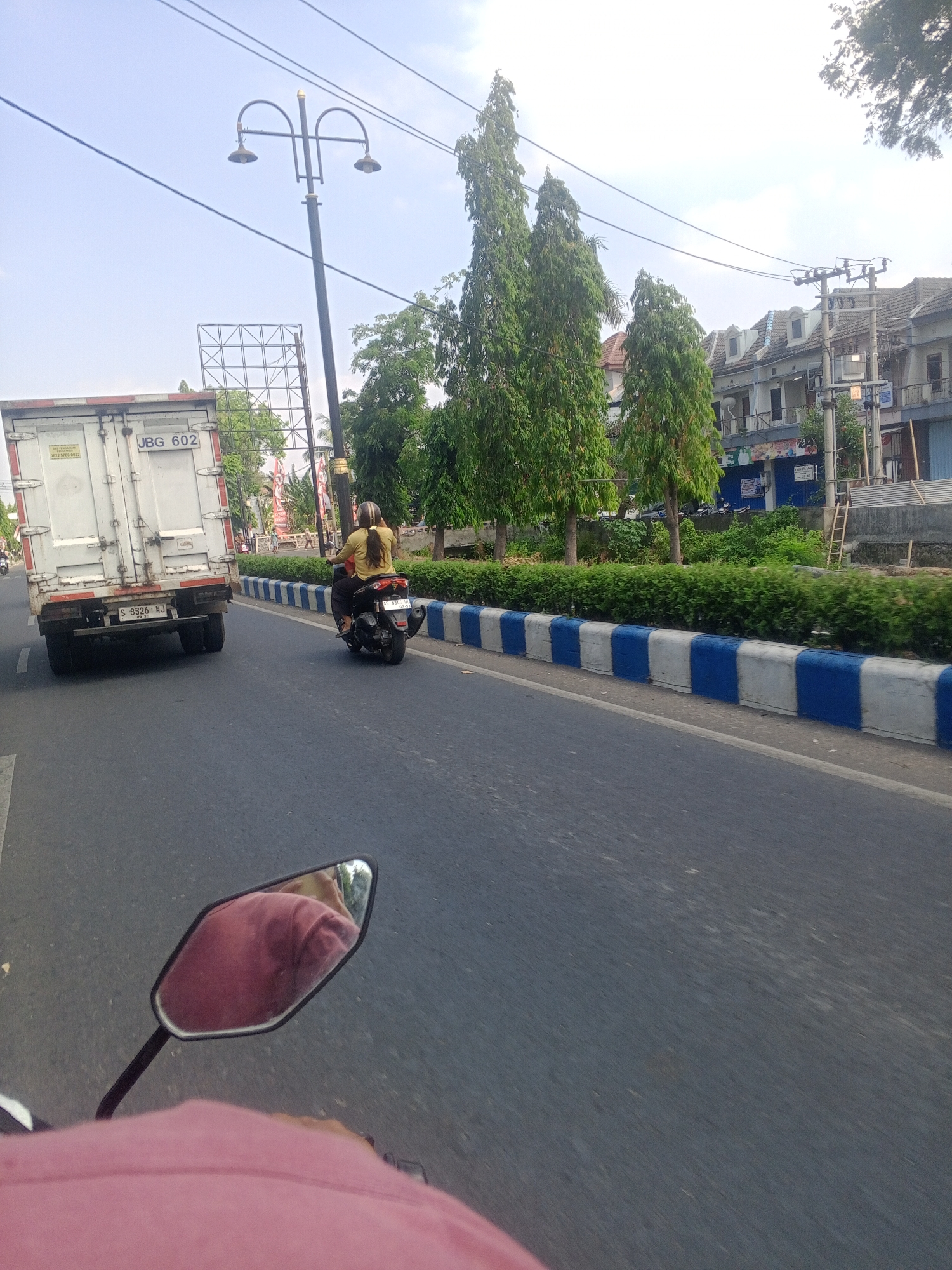 This image captures a street scene in Tonatan, Indonesia, likely during the daytime given the bright sky. The perspective is from a vehicle, possibly a motorcycle, as indicated by the handlebar and mirror visible in the foreground.

On the road, a white truck with the license plate "JBG 602" is in motion. To the right of the truck, a person on a motorcycle, wearing a yellow top and a helmet, is also driving. The road is paved and appears to be a two-lane highway with a median barrier painted in alternating blue and white stripes.

The environment includes a row of tall, slender trees lining the median. Beyond the trees, buildings with tiled roofs are visible, suggesting a commercial or residential area. A street lamp with two lights stands on the left side of the road. Electrical wires are strung across the sky, a common sight in urban Indonesian landscapes. The overall atmosphere suggests a typical day on a busy road in Indonesia.