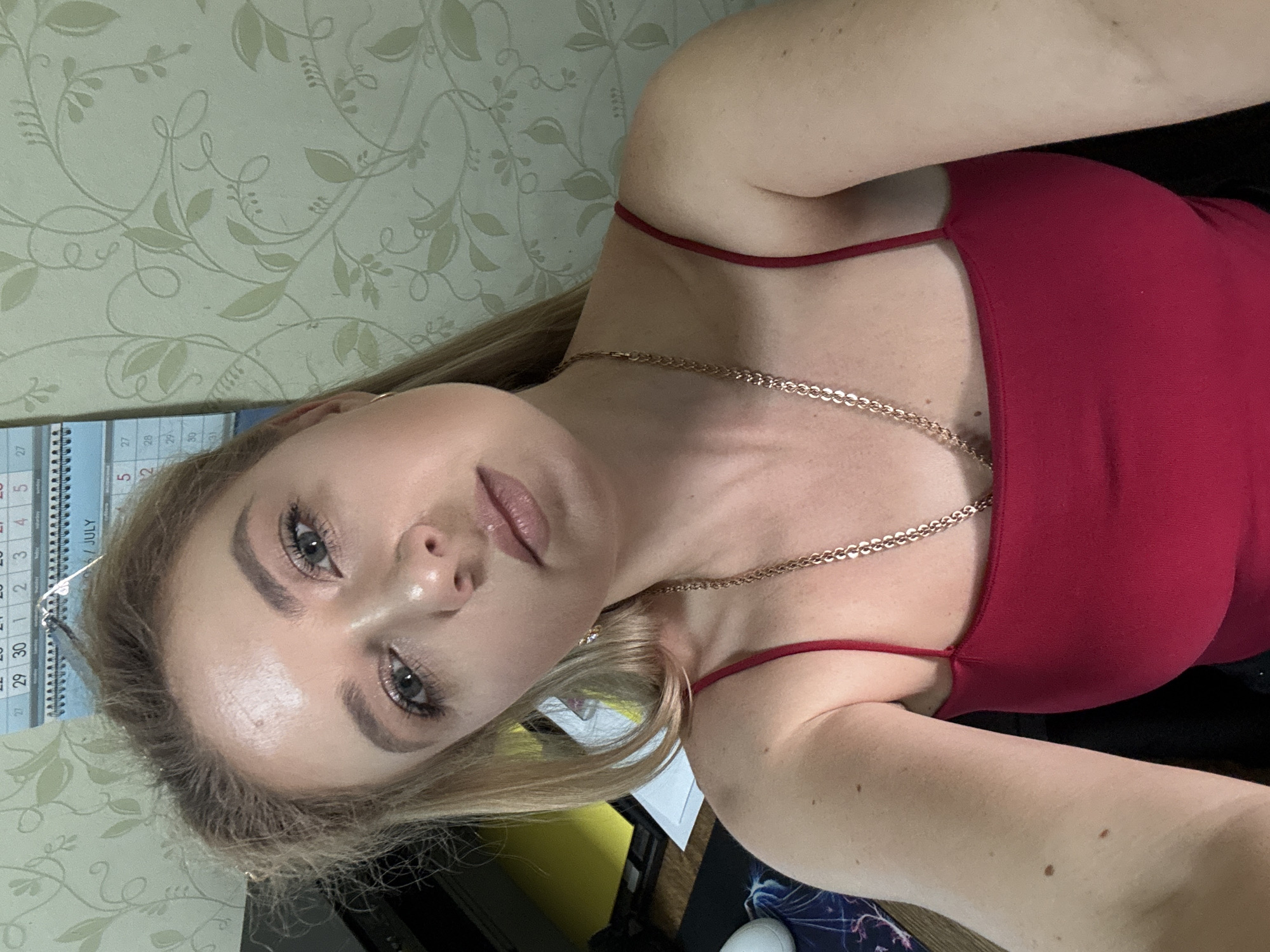 A blonde woman with light skin and makeup is looking at the camera, leaning her head to the right. She is wearing a red spaghetti strap top and a gold chain necklace. The background consists of a light green floral wallpaper and a calendar. The lighting is natural.