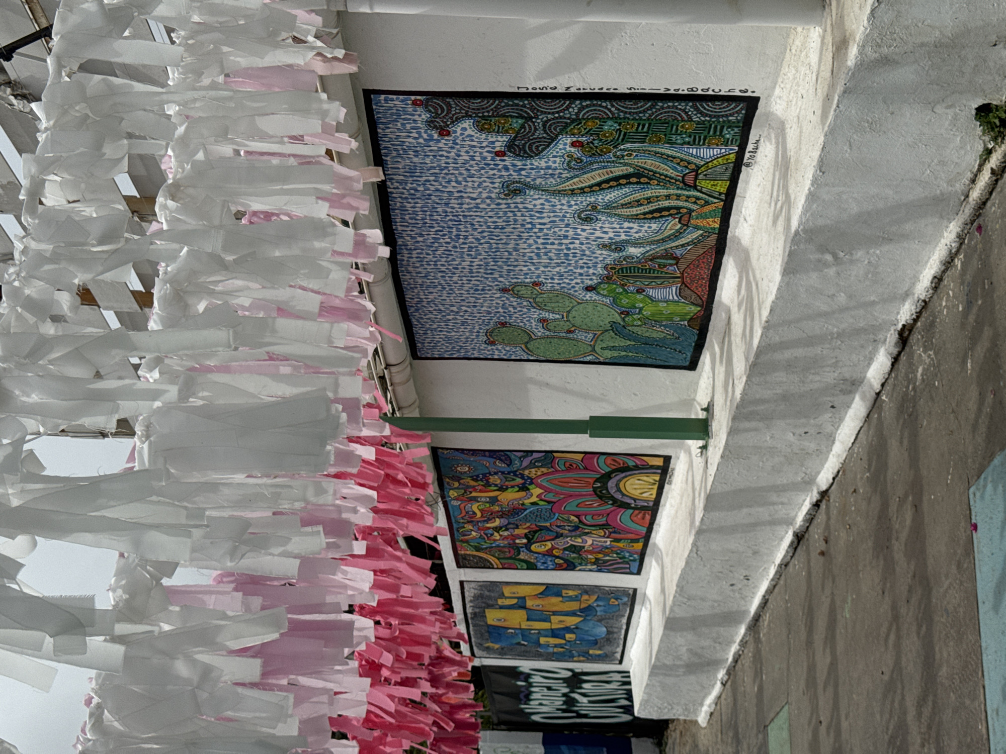 The image shows a street scene, likely in Pampatar, Venezuela, judging by the "Manjeiro Cultura" sign visible towards the bottom. The focus is on a building's exterior wall adorned with artwork. Three framed art pieces are stacked vertically: the top one depicts a stylized landscape with cacti and swirling patterns in blue, green, and brown; the middle one features a colorful, mandala-like design; the bottom one shows a geometric pattern in blue and yellow. White and pink paper decorations hang along the wall, obscuring the view and extending towards the left edge of the frame. The wall is painted white, with a concrete sidewalk visible along its base. A green metal bar extends horizontally above the artwork. The overall impression is of a vibrant, decorated street scene.