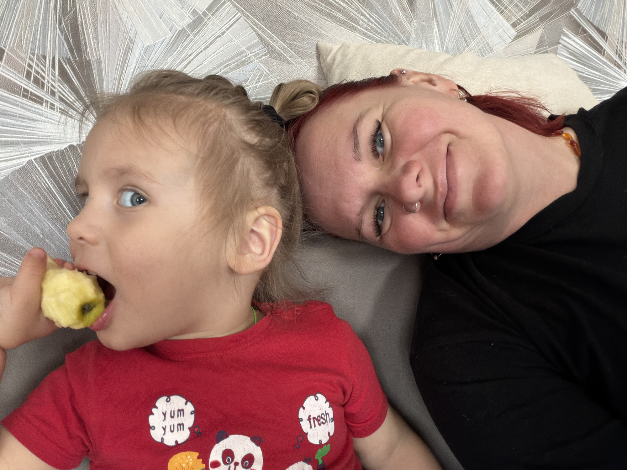 A photograph taken indoors shows a woman and a young child lying on a gray surface. The child, on the left, is fair-skinned with blonde hair pulled back in a small ponytail. The child is wearing a red shirt with panda bear designs and is biting into a peeled fruit, possibly an apple. The woman, on the right, is fair-skinned with red hair and is wearing a black top. A light-colored pillow is positioned behind her head. The background features a wall with a geometric pattern. The location is Chaikovsky, Russia.