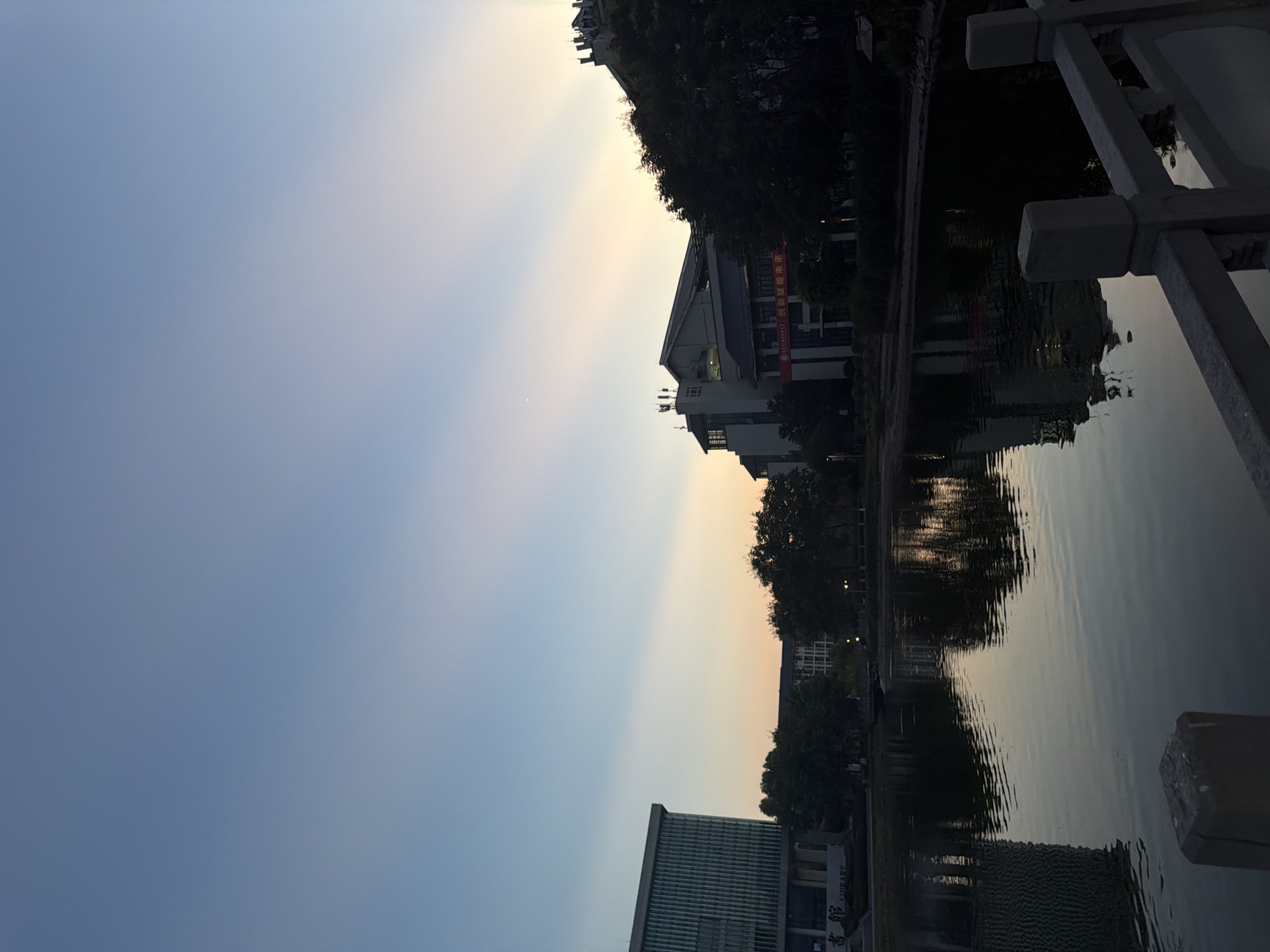 The image depicts a serene twilight scene at the Jiangjin Library, located in Jiangjin, China. A calm lake reflects the soft hues of the sky, which transitions from a deep blue at the top to a pale yellow and orange near the horizon, with visible crepuscular rays emanating from a hidden light source. On the left side of the frame, a modern, multi-story building, identifiable as the library by its sign which reads "图书馆" (Library), stands prominently. On the right, a darker, more traditional building with multiple antennas on its roof is partially obscured by silhouetted trees. The foreground features a stone railing, part of a bridge or walkway, overlooking the lake. There are no people visible in the scene.