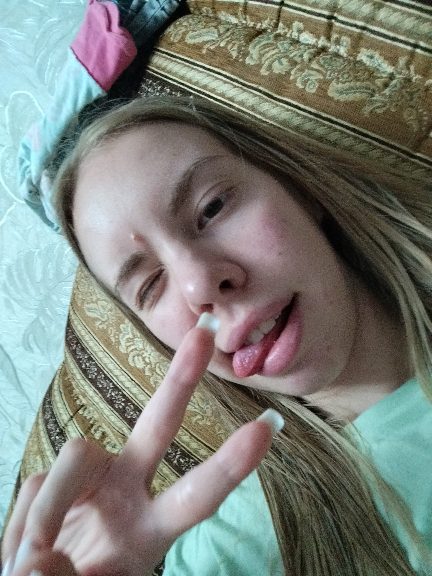 A young Caucasian woman with long blonde hair is lying down and taking a selfie. She is wearing a light blue t-shirt and has her tongue sticking out of her mouth, with her left hand raised and making a peace sign. The background appears to be a bed with a patterned comforter and a light blue sheet. The lighting suggests an indoor setting, possibly a bedroom, with no specific city or location details visible.
