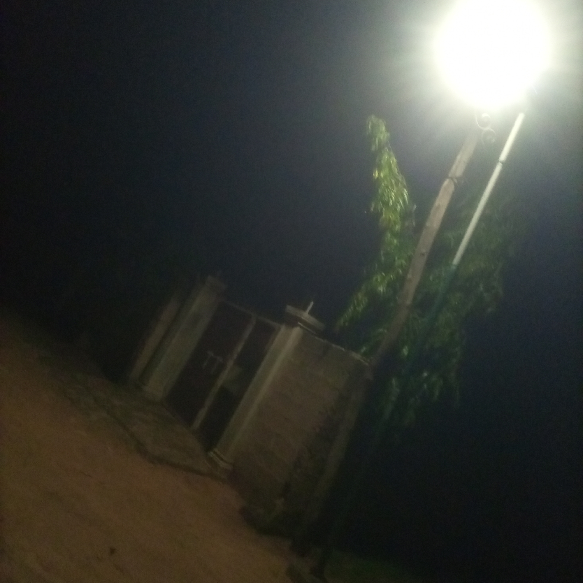 The image depicts a street light illuminating a dark environment at night. The light source is a bright, white lamp at the top of a pole. To the left of the pole, a tree with green foliage is visible, extending upwards into the darkness. Below the tree and to the left, a building with a rectangular structure and darker openings is partially visible. The ground in the foreground appears to be dirt or gravel. The overall scene is dimly lit, with the bright light of the street lamp creating a strong contrast against the dark surroundings. There are no people visible in the image.