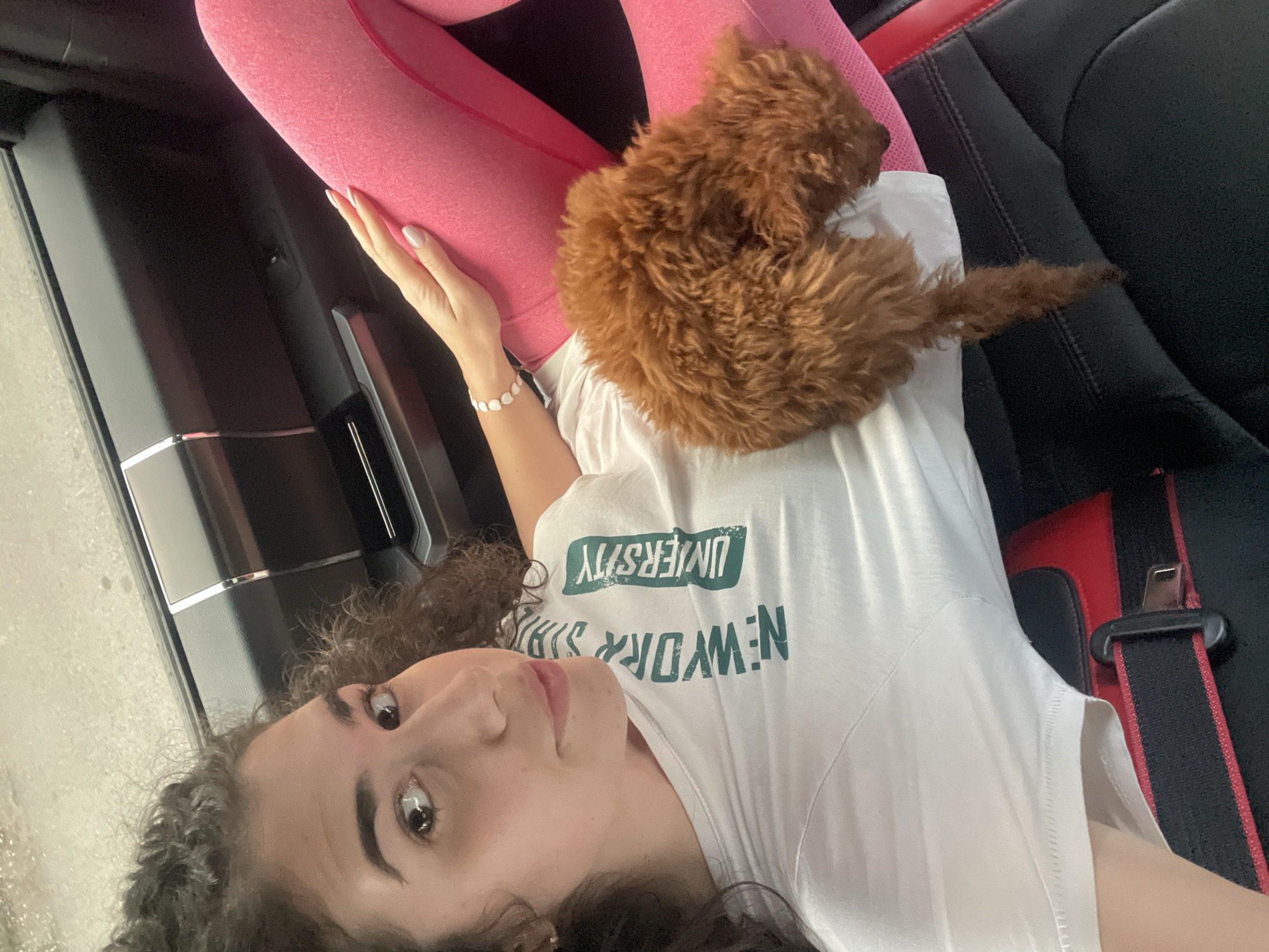 This media file captures a close-up, high-angle shot of a young woman and a small dog resting inside what appears to be a car in Sofia, Bulgaria. The image is taken from above, with the woman lying on her back, looking up towards the camera.
The primary subjects are a woman with dark, curly hair and a small, fluffy, reddish-brown dog. The woman is wearing pink leggings and a white t-shirt with green text. Her left arm is visible, adorned with a delicate bracelet featuring light-colored, possibly pearlescent, beads. Her fingernails are neatly manicured with a light, neutral polish. She has an open-mouthed, relaxed expression, and her eyes are directed towards the viewer.
The small dog, which appears to be a puppy of a poodle or similar breed, is curled up and resting on the woman's chest/lap area. Its tail is visible extending towards the right, against a dark car seat.
The setting is an automobile interior, evidenced by parts of a black car seat with red accents on the right, and a dark car door panel with metallic trim visible on the left. A black and red seatbelt is also visible near the woman's right shoulder. The lighting suggests it is daytime, with soft, indirect illumination.
Visible text on the woman's white t-shirt, oriented upside down from the viewer's perspective, clearly reads "UNIVERSITY" in green letters, with "NEW YORK" partially obscured by her hair.
The overall mood is calm and affectionate, depicting a relaxed moment between the woman and her canine companion.