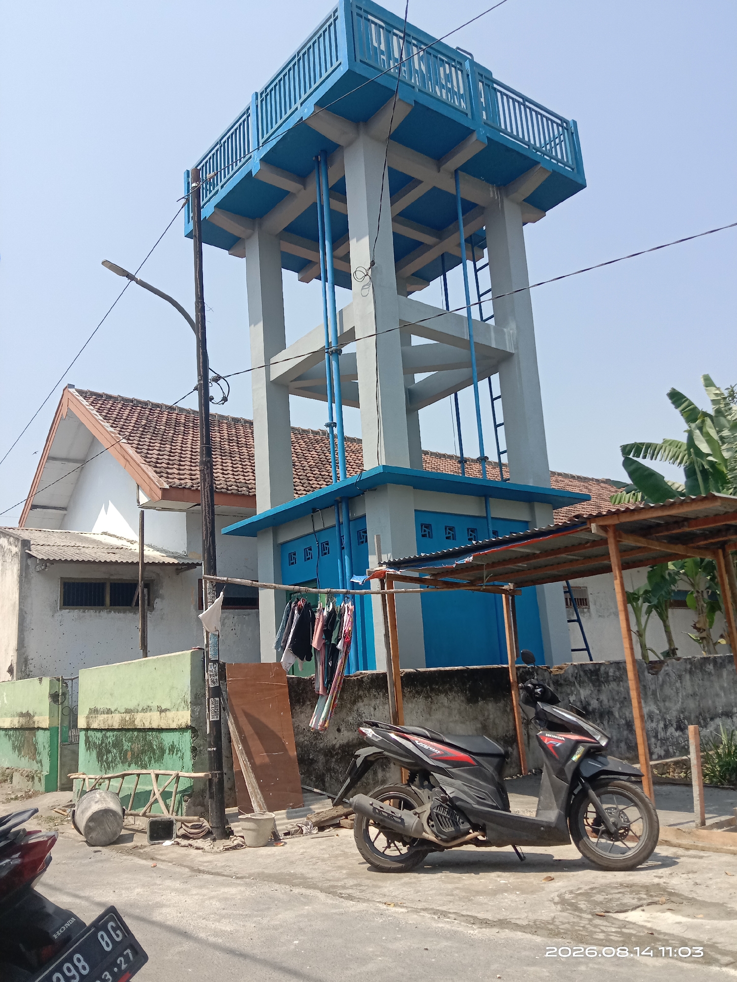 The image depicts a scene in Blimbing, Indonesia, featuring a prominent blue and gray water tower. The water tower has a multi-level structure with a blue railing on the uppermost platform and blue pipes running down its sides. Adjacent to the water tower is a white building with a red-tiled roof and several blue-gridded windows. In front of these structures, a black motorcycle is parked on a paved surface. To the left of the motorcycle, a wooden ladder leans against a green and white wall. A clothesline with various garments is strung between two poles in front of the blue-gridded windows. A street light is visible to the left of the water tower. The ground is a concrete pathway, with some debris and a large concrete cylinder visible. The sky is clear and blue. A timestamp in the lower right corner reads "2026.08.14 11:03".