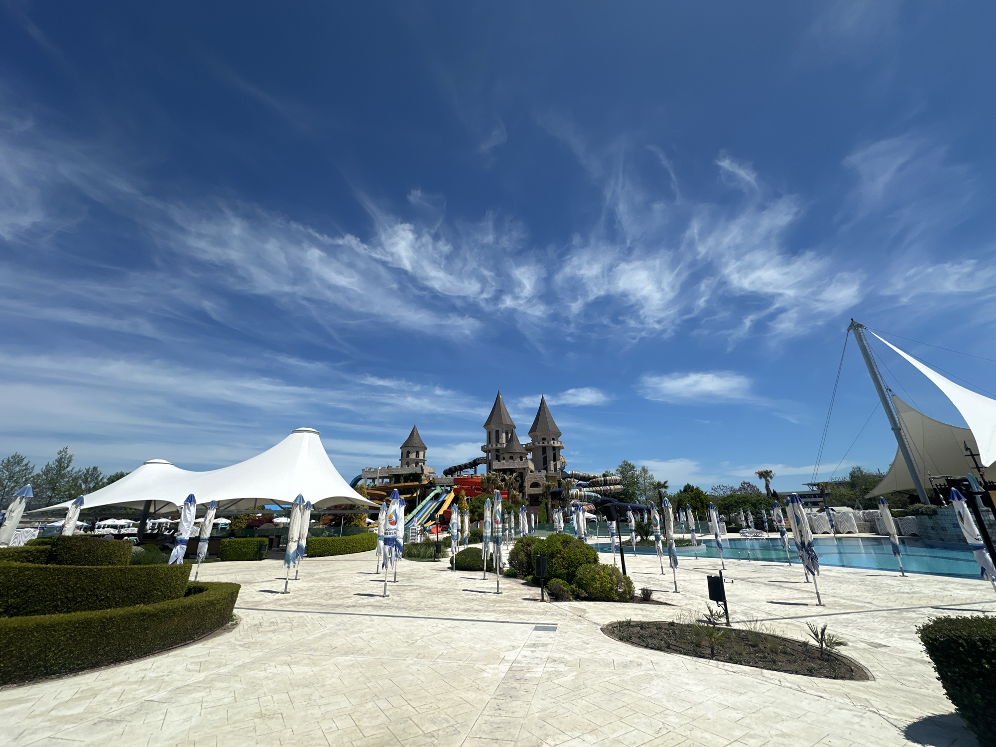 The image depicts a sunny day at the Aqua Paradise water park in Ravda, Bulgaria. The scene is generally deserted. In the background is a castle-themed structure with multiple water slides. Several large white sun shades provide cover. Many folded umbrellas stand in their bases throughout the area. A swimming pool is visible on the right. The ground is paved with patterned, light-colored stone. The sky is blue with scattered wispy clouds. Green shrubbery is present in various locations.