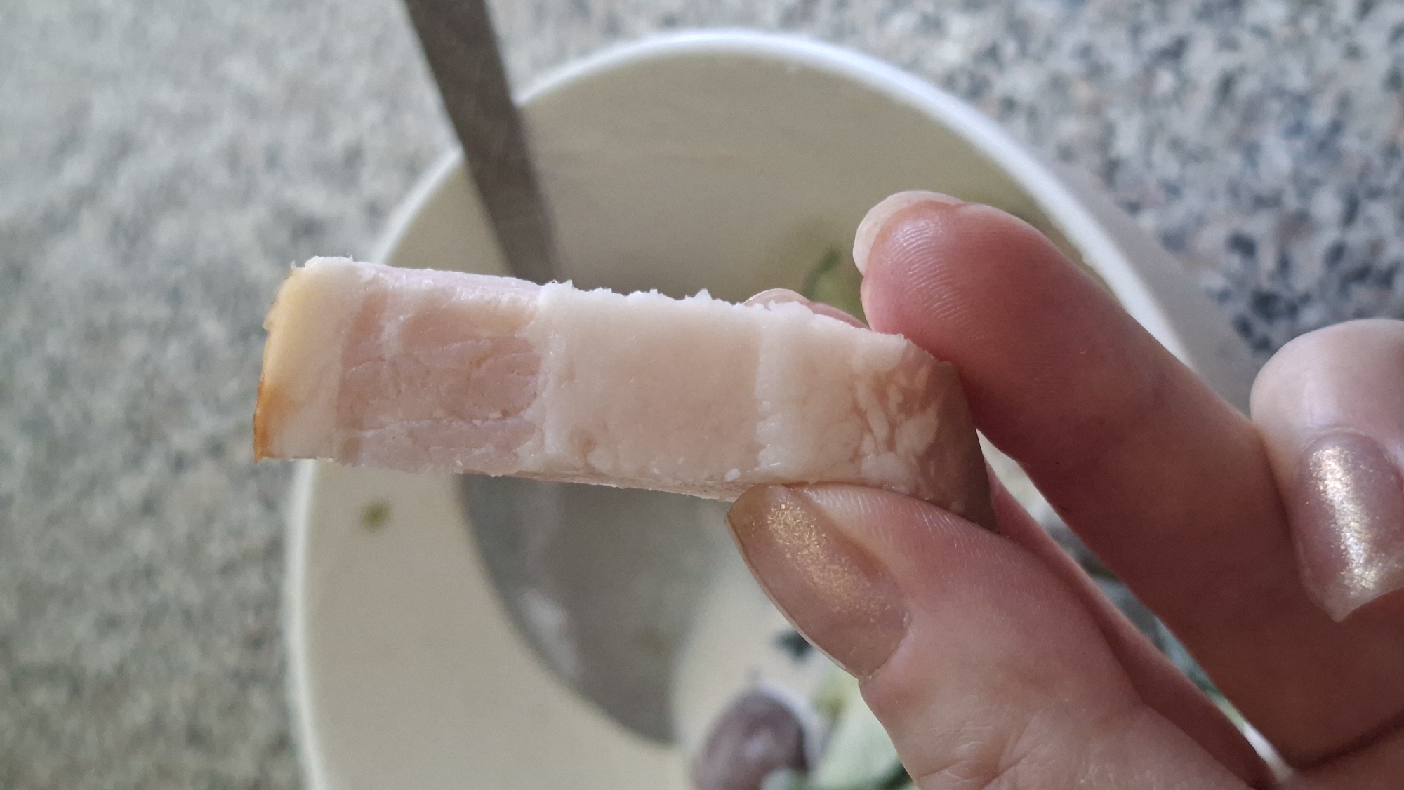 A close-up shot shows a person holding a piece of cured bacon between their thumb and index finger. The bacon is rectangular and has distinct layers of white fat and pinkish meat. The person's fingernails are manicured with a shimmery, light-colored polish. In the background, out of focus, is a white bowl containing what appears to be a creamy soup or salad with some green ingredients. A silver spoon is partially visible in the bowl. The surface on which the bowl rests appears to be a speckled countertop. The image is taken from a slightly upward angle. The location is not discernible from the image itself.