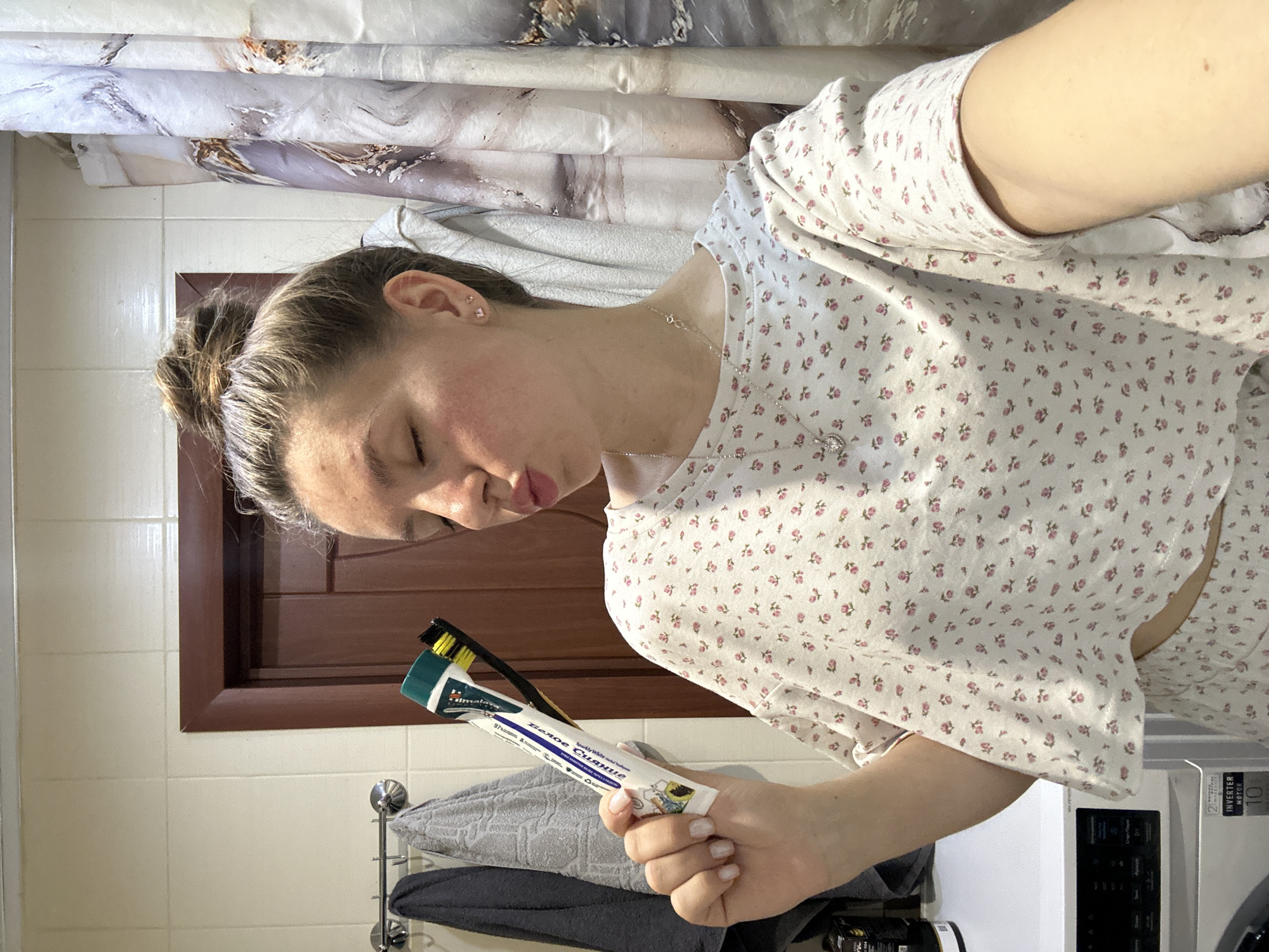 A woman is standing in a bathroom. She is wearing a white shirt with small pink flowers and a necklace. In her hand, she holds a toothpaste tube labeled in Russian and English, "Sparkly White." A toothbrush with a black handle and yellow bristles is also in her hand. Behind her, there is a bathroom shower curtain, a brown wooden door, a towel rack with towels, and a washing machine. The location is likely not in Klin, Russia, as there is no evidence of that in the image.
