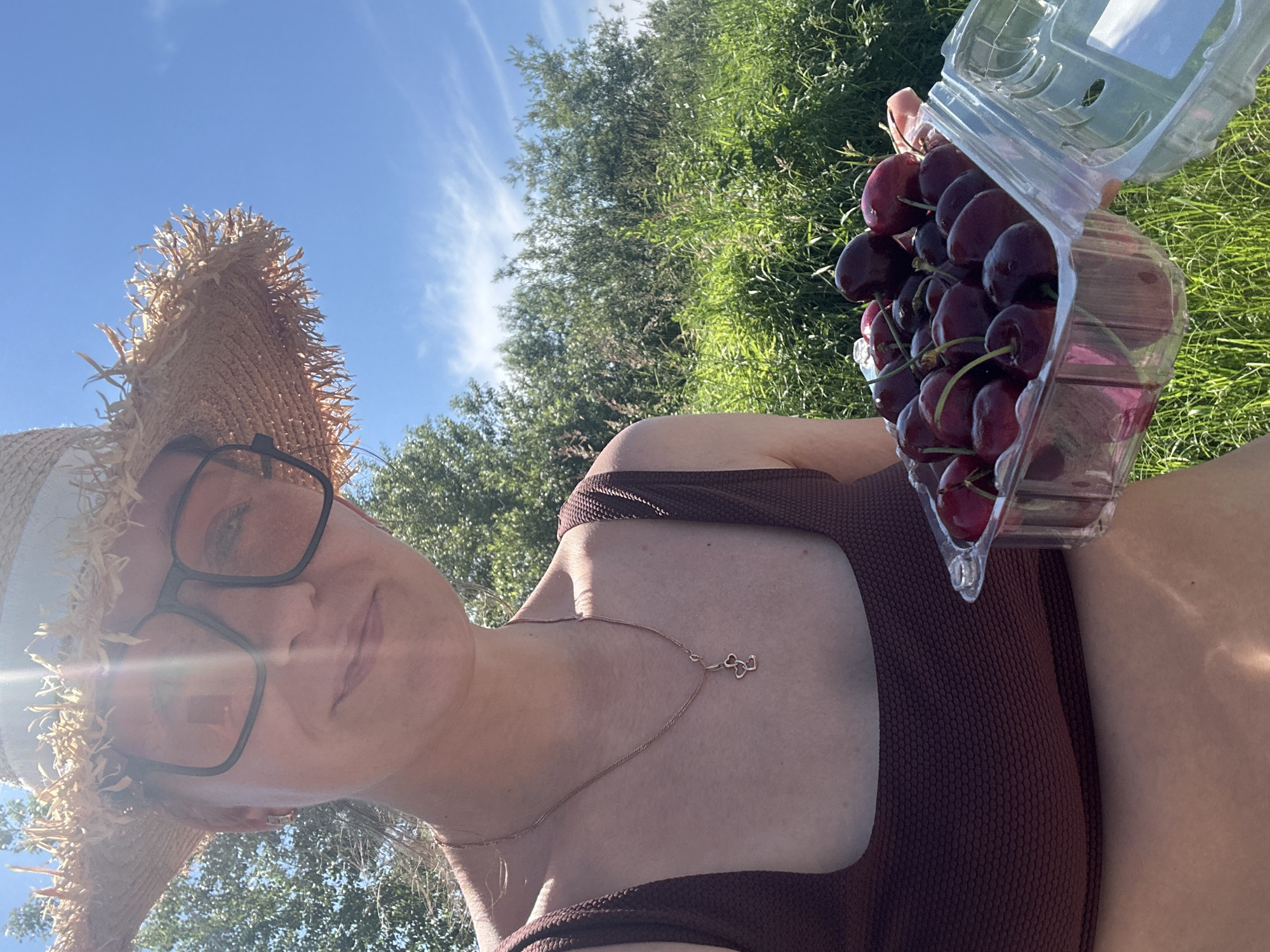A person wearing a straw hat and glasses holds a clear plastic container filled with dark red cherries. The person is wearing a brown textured swimsuit top. The background consists of lush green foliage and a bright blue sky with scattered white clouds. The scene appears to be outdoors in a natural setting.