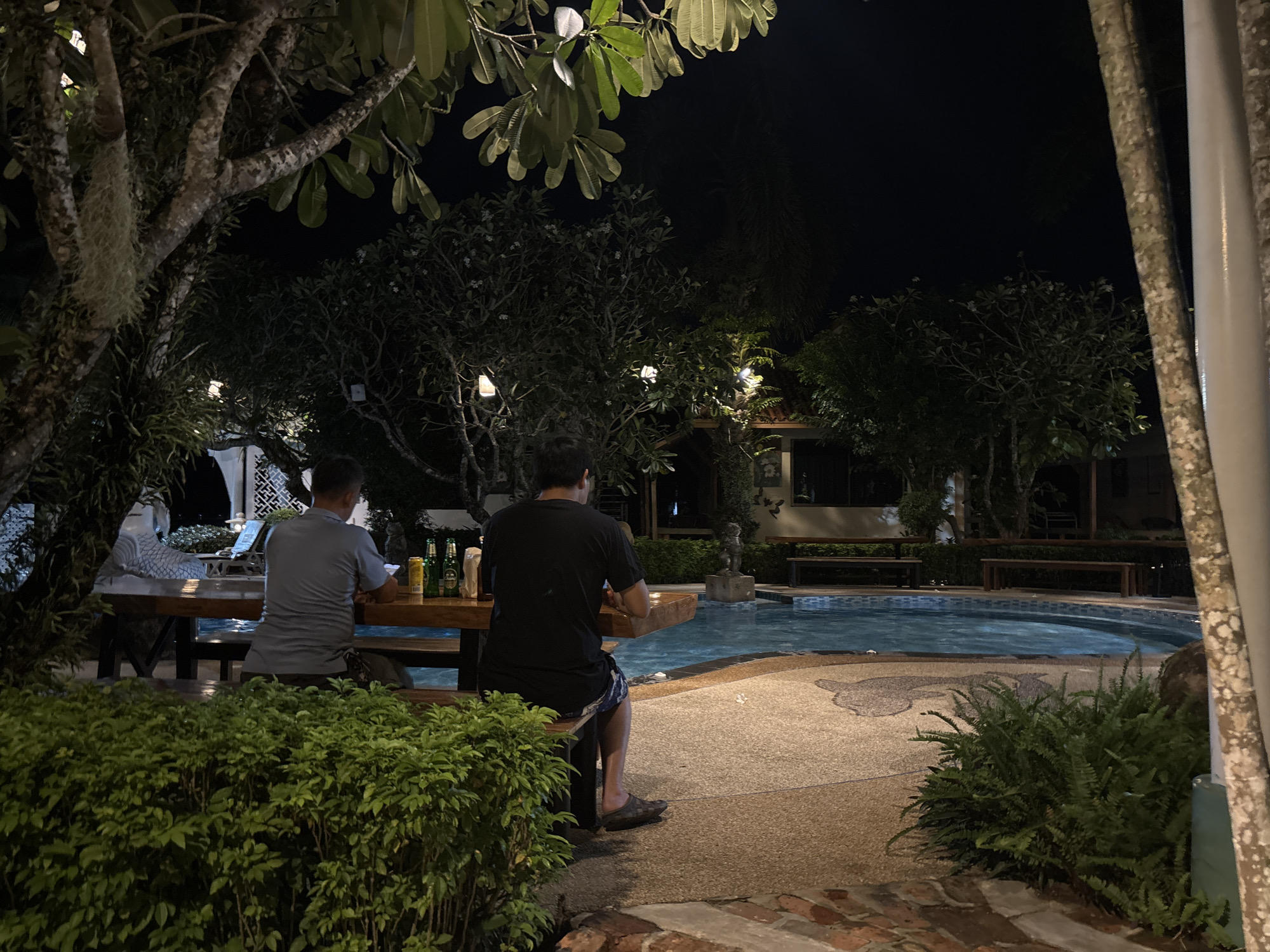 At night, two men are seated at a wooden table by a swimming pool in a resort located in Chalong, Thailand. The man on the left, wearing a gray polo shirt, faces away from the camera and is seated at the far end of the table. On the table, there are several bottles of beer and a can. The man on the right, wearing a black t-shirt and patterned shorts, is closer to the camera and also faces away. He is seated on a bench at the table. The swimming pool is illuminated and appears to be empty. Lush green bushes are in the foreground, and trees with hanging lights are visible in the background. The overall setting is an outdoor dining area of a resort, illuminated by ambient lighting.